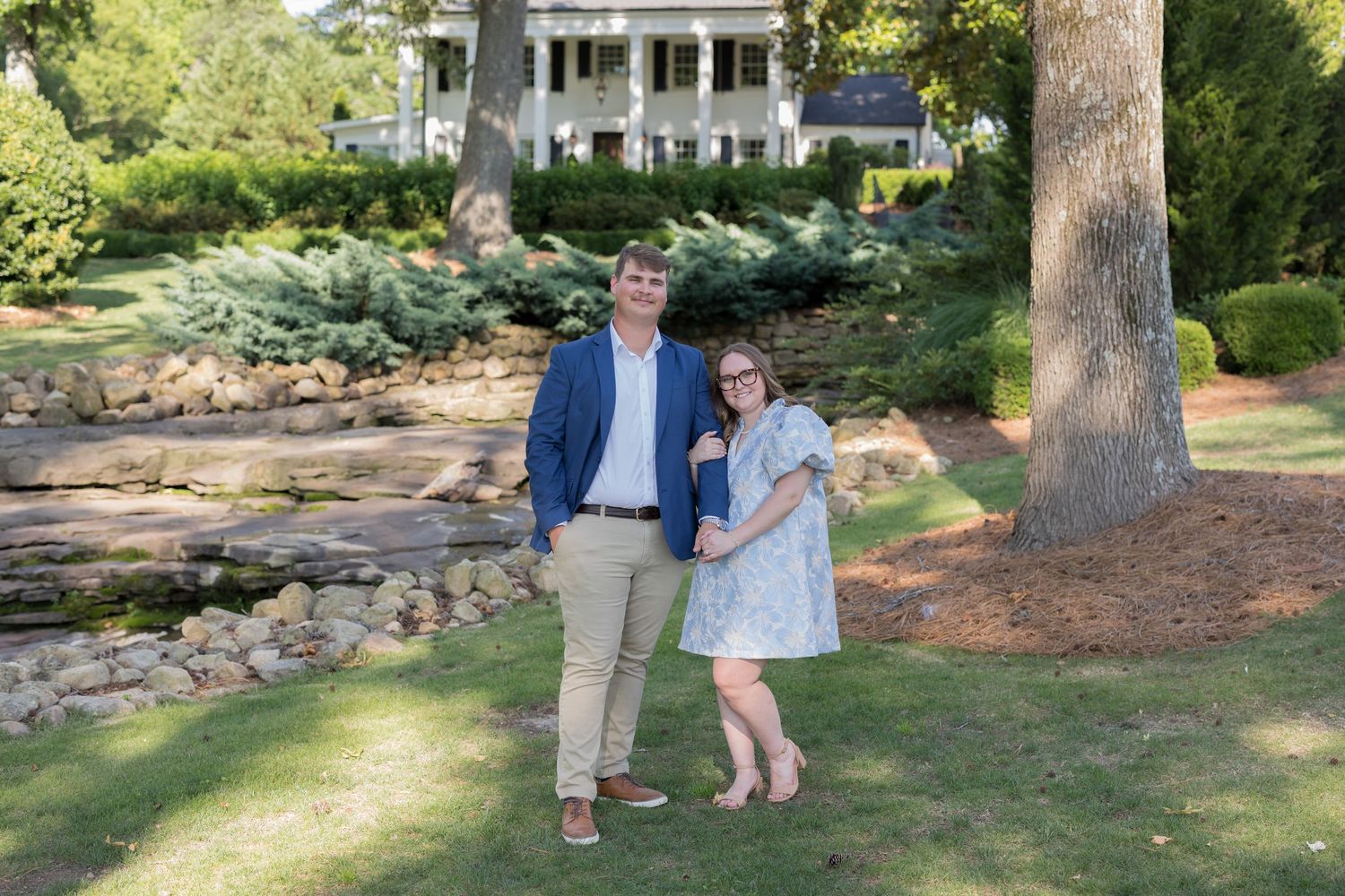 Couple posing together in front of a white Southern mansion surrounded by lush greenery and stone landscaping.