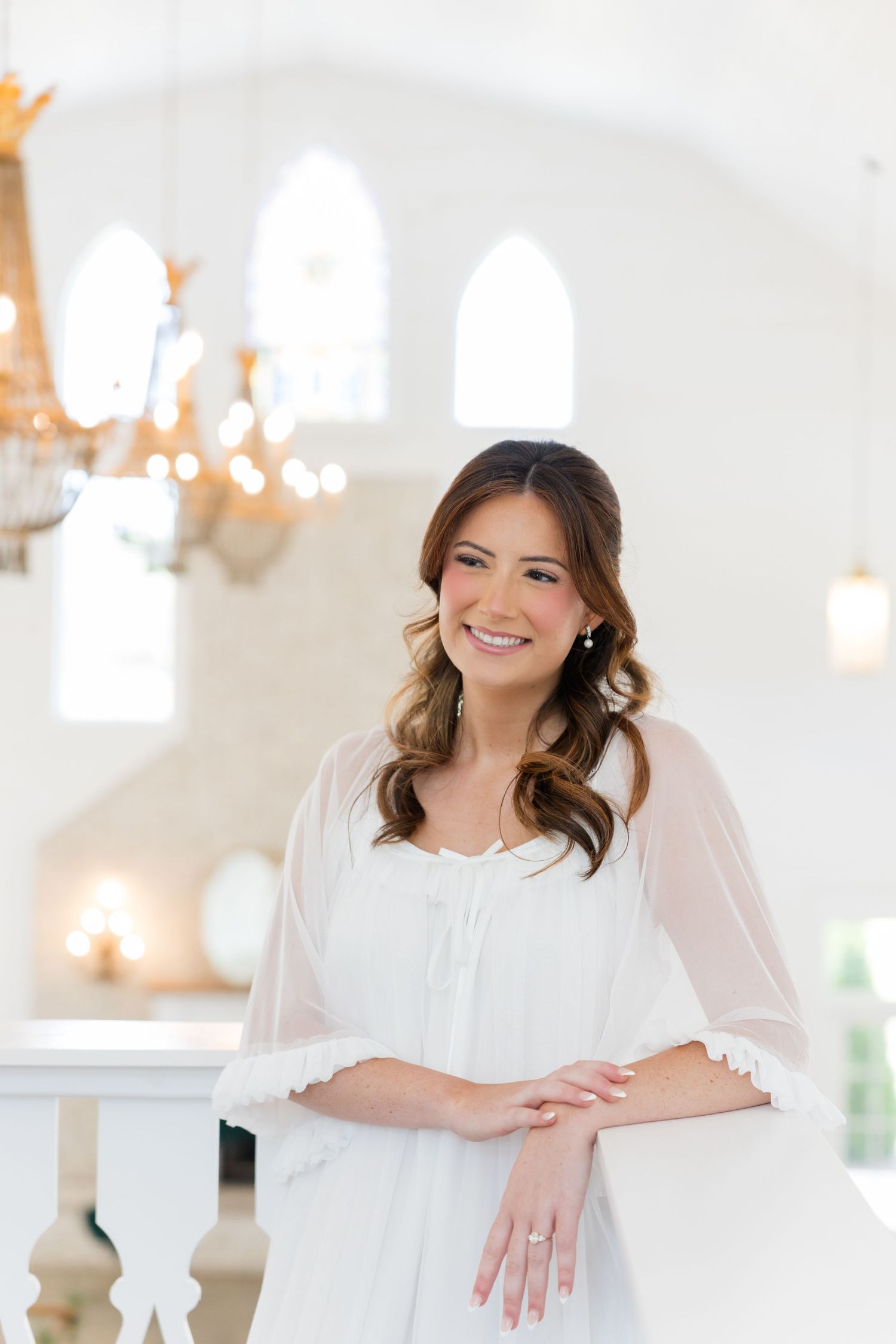 Smiling bride in white dress and sheer cape leans on balcony railing in elegant white venue with chandelier.