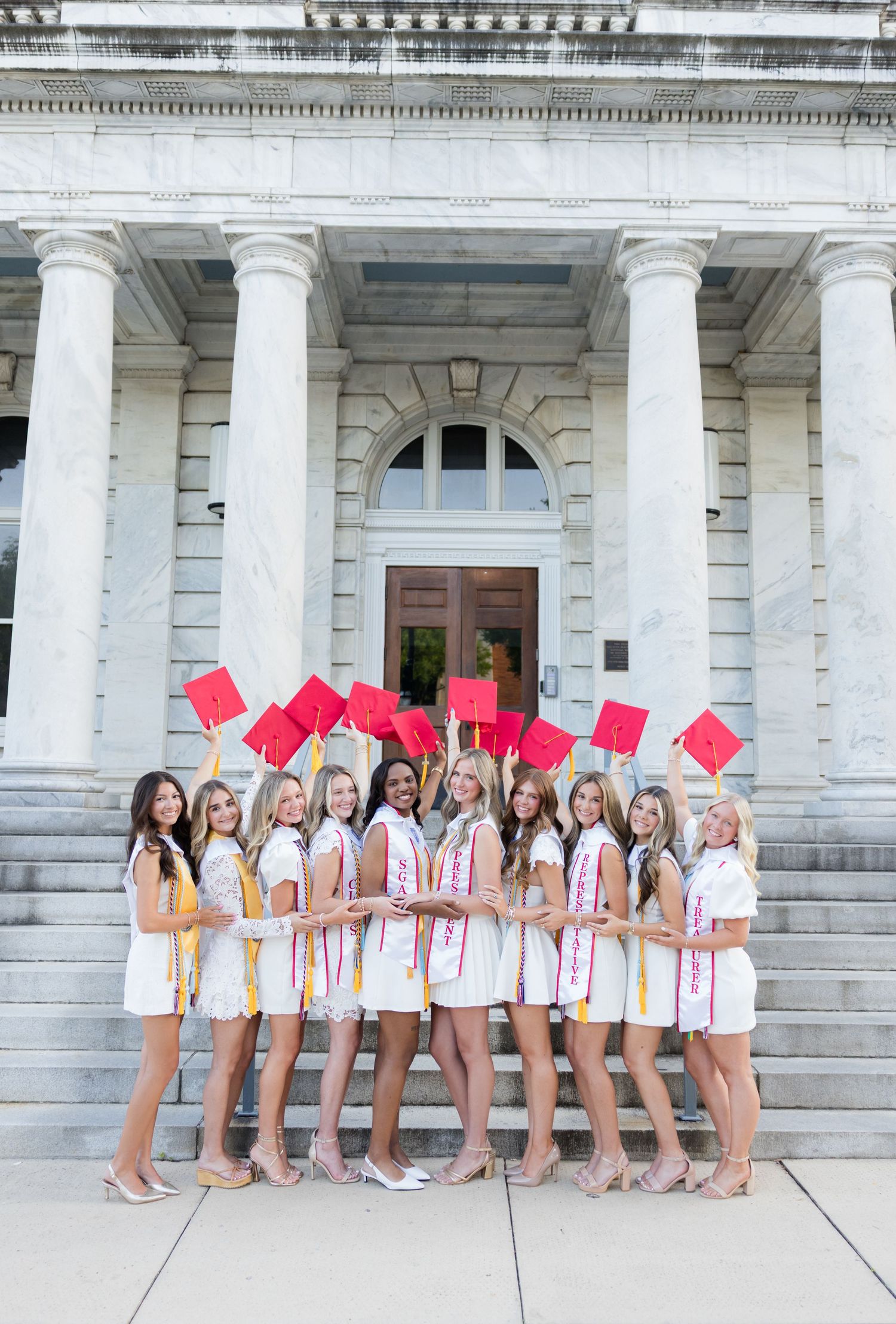 Group of female graduates in white dresses holding red caps in front of a grand columned building.