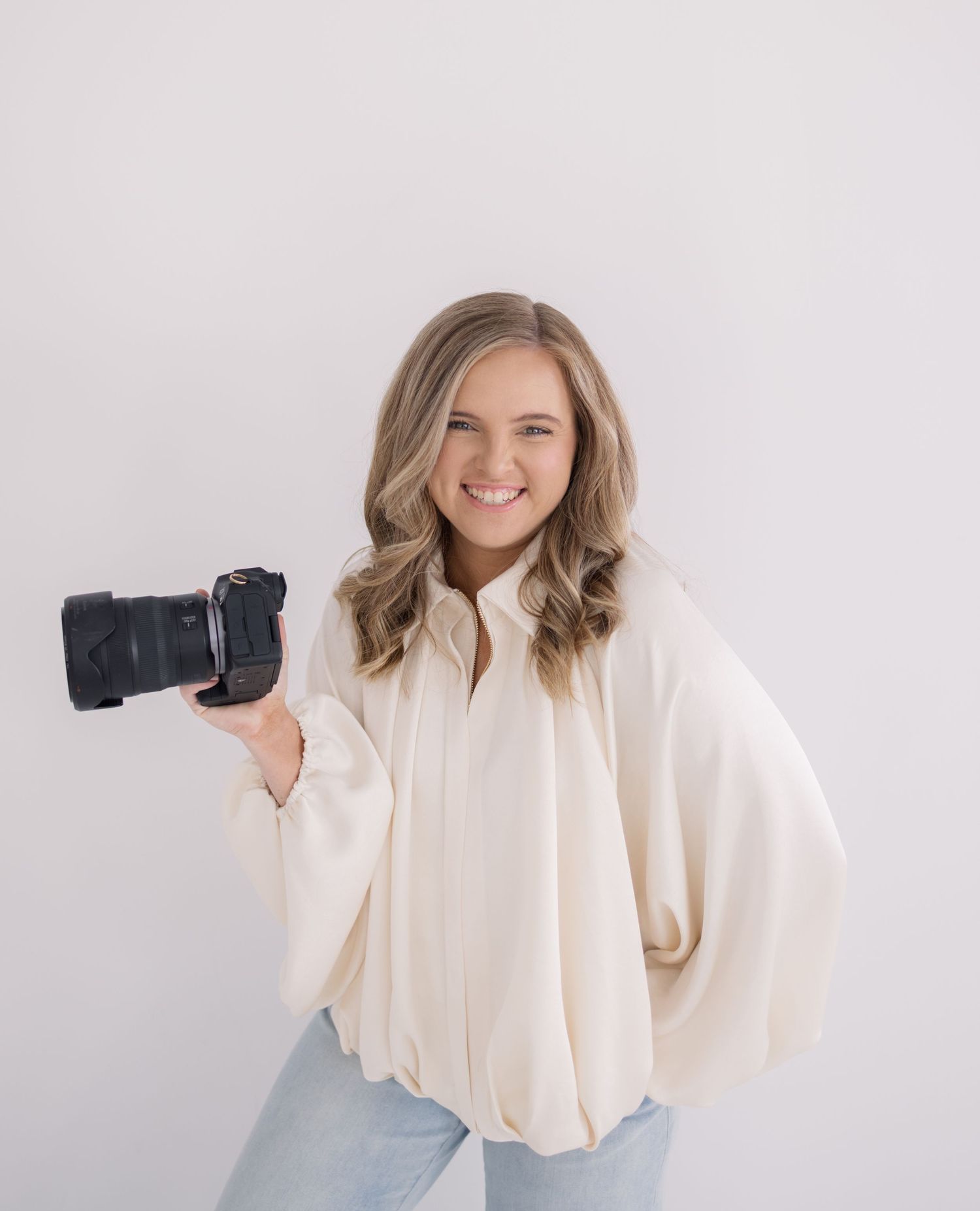 Smiling female photographer in cream oversized blouse holds a DSLR camera against a clean white backdrop.