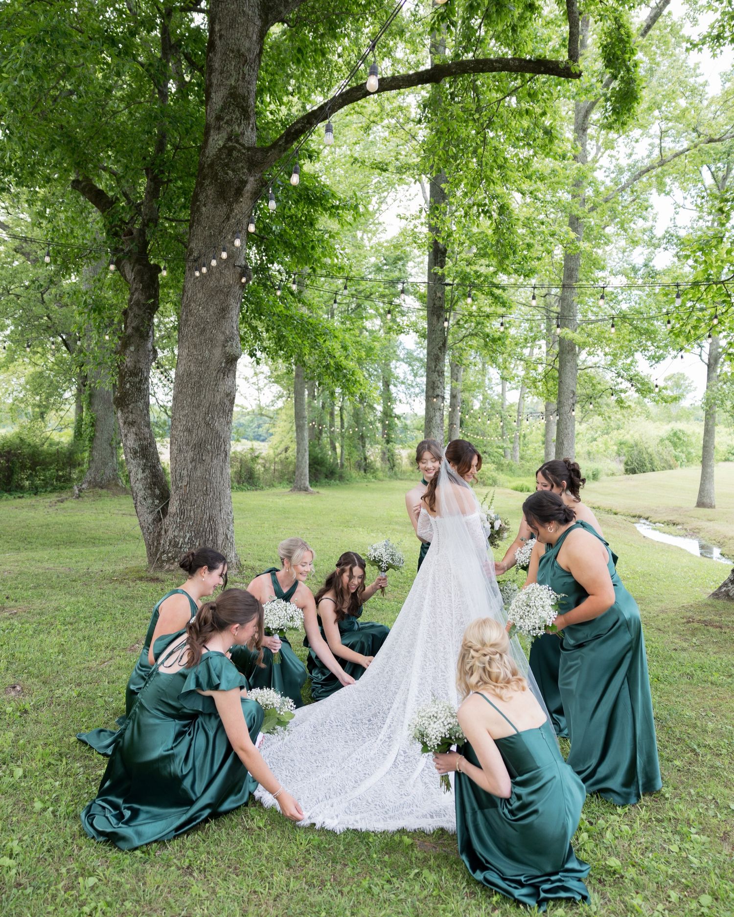 Bride in white lace gown surrounded by bridesmaids in emerald green satin dresses arranging her train outdoors.