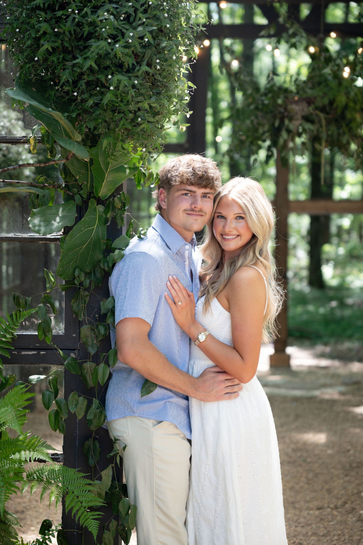 A smiling couple poses outdoors near lush greenery, the woman in white dress and man in blue shirt embracing warmly.