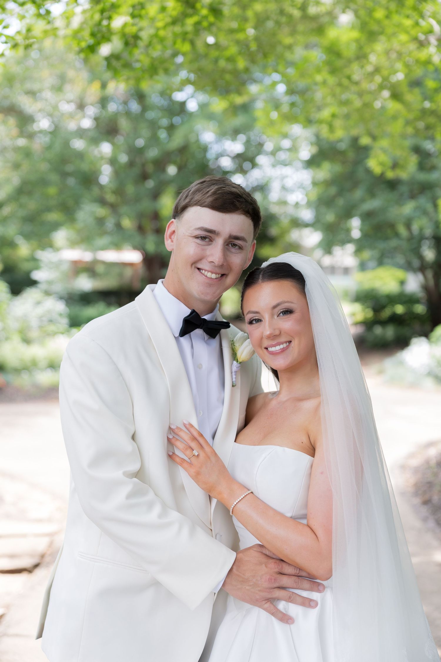 Happy newlyweds pose outdoors, groom in white tuxedo and bride in strapless gown with veil, surrounded by lush greenery.