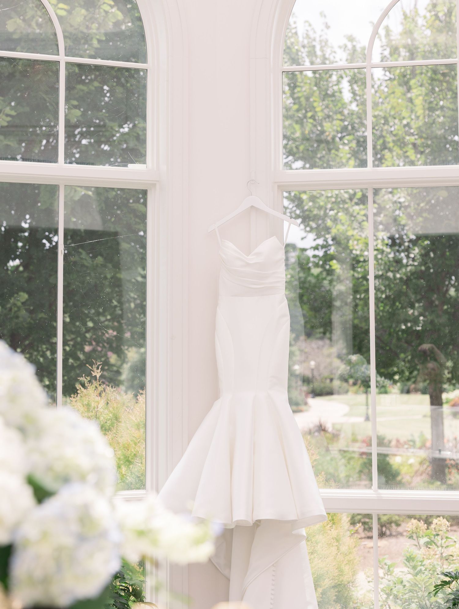 Elegant strapless mermaid wedding dress hanging in a sunlit arched window with garden views.