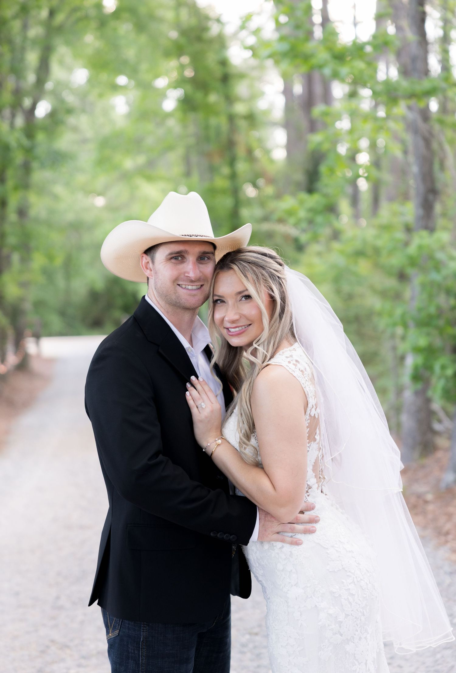 Bride in lace gown and veil embraces groom in black suit and white cowboy hat on a tree-lined path.