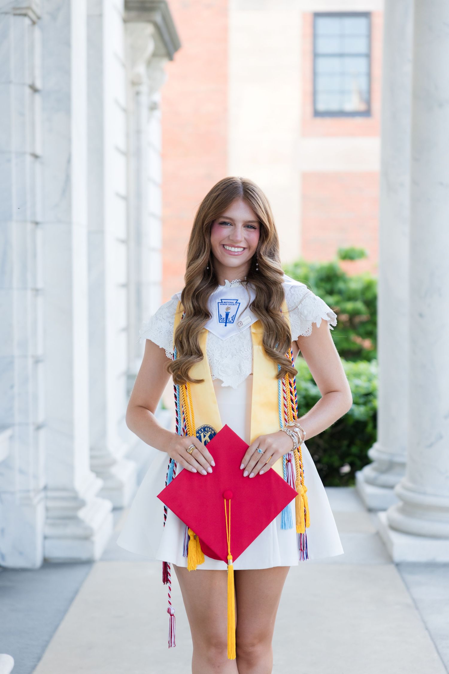 Smiling graduate in white dress holding red cap with colorful honor cords posed between white columns.