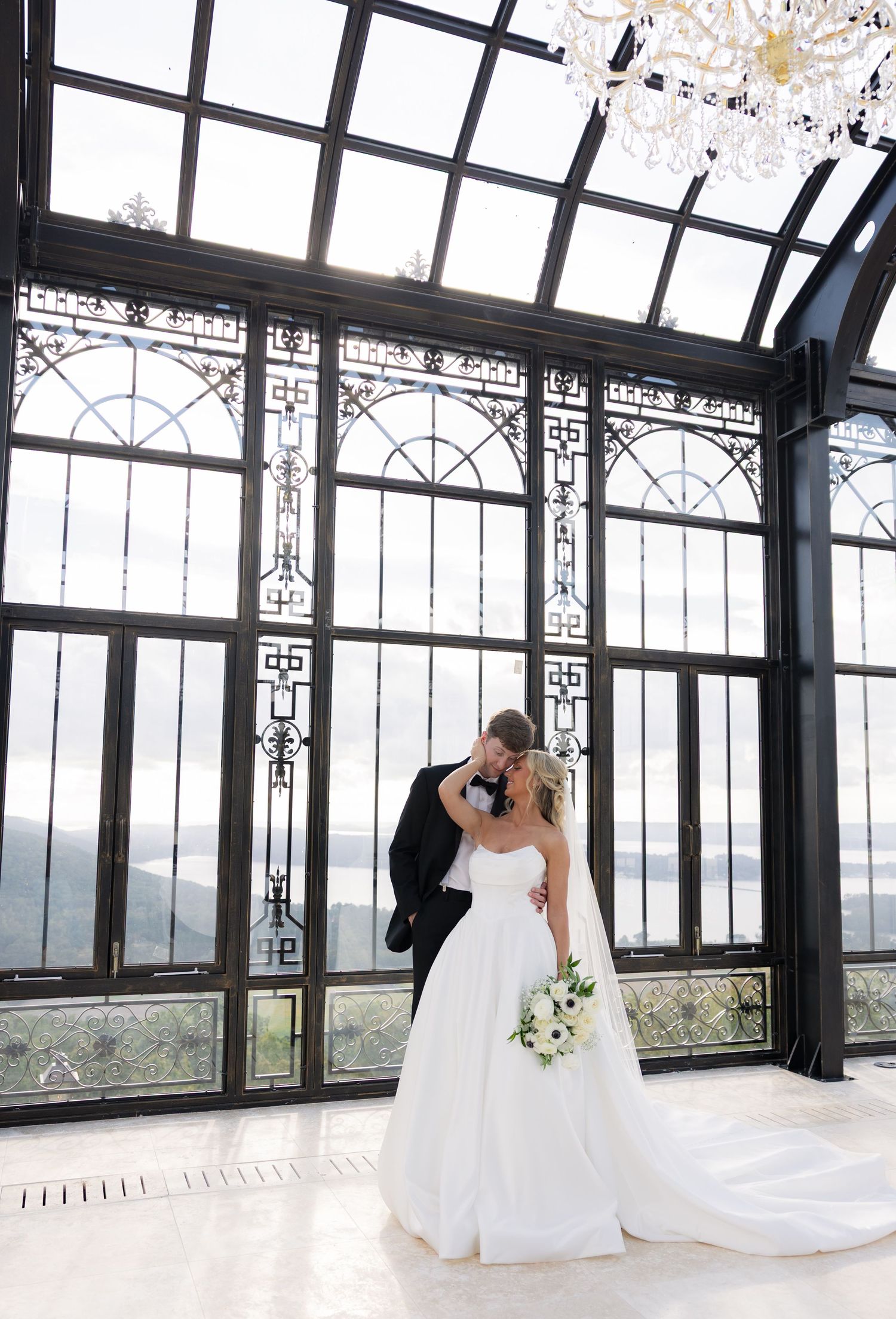 Bride and groom share a kiss in an elegant glass conservatory with ornate black frames and a crystal chandelier.