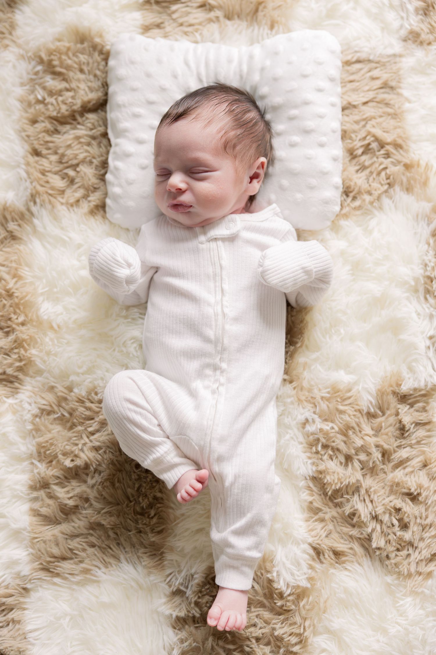 Peaceful newborn baby sleeping in white onesie on fluffy beige rug with small white pillow.