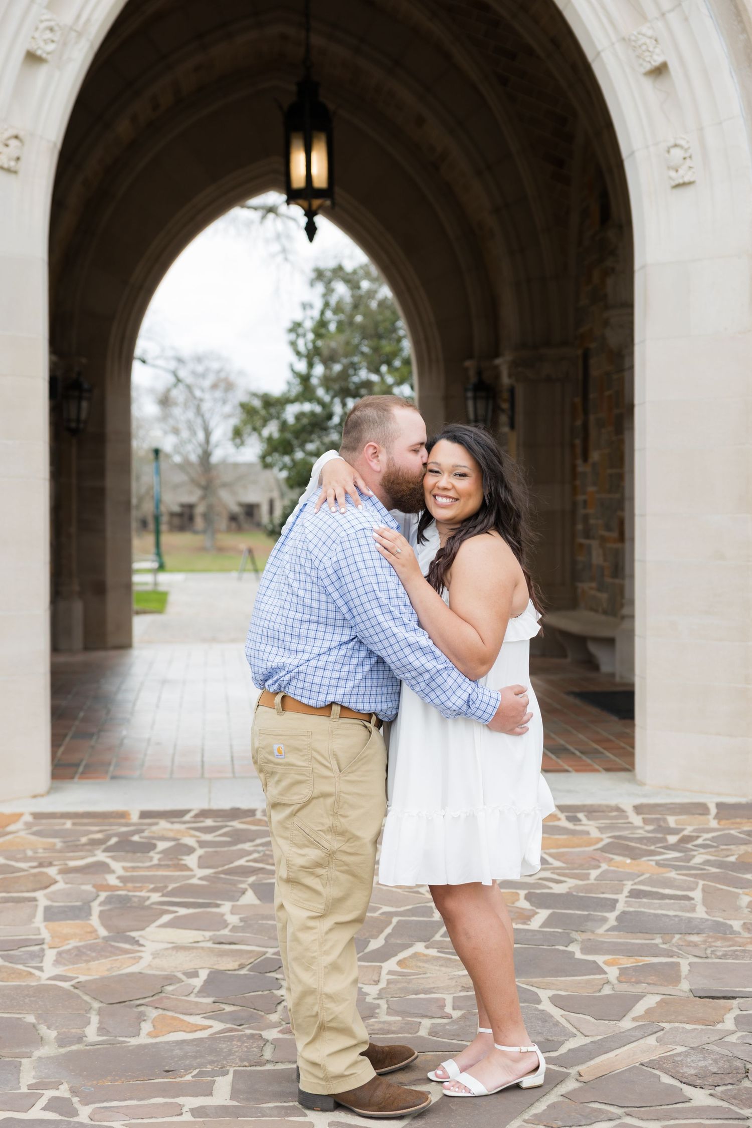Couple embracing and smiling under a stone arch at an outdoor venue, woman in white dress, man in blue plaid shirt.