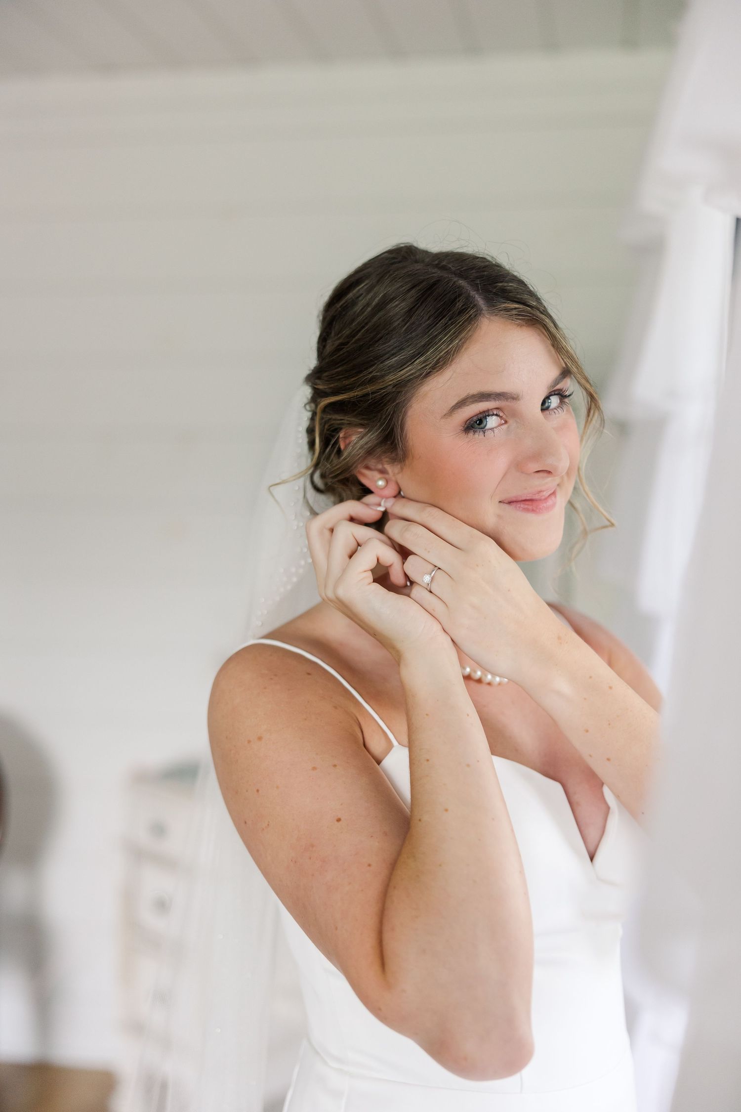 Bride in white dress putting on earrings with pearl bracelet and engagement ring, smiling softly in bright room.