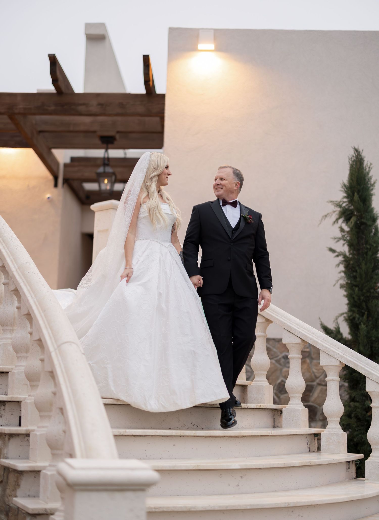 Bride in flowing white gown and veil walks hand-in-hand with groom in black tuxedo down elegant stone staircase.