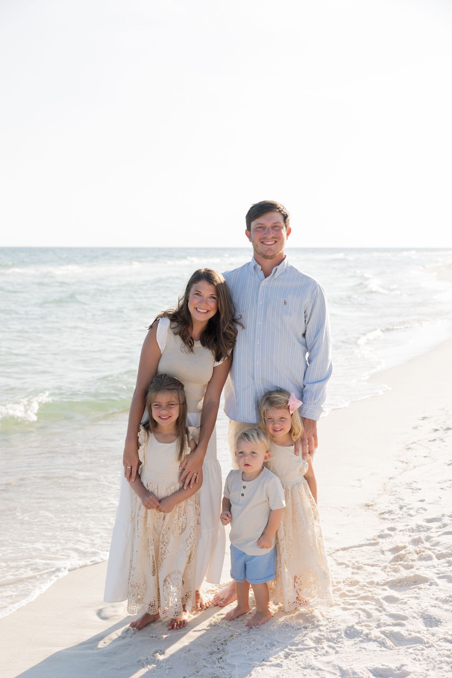 Happy family of five posing together on a sunny beach with turquoise ocean waves in the background.