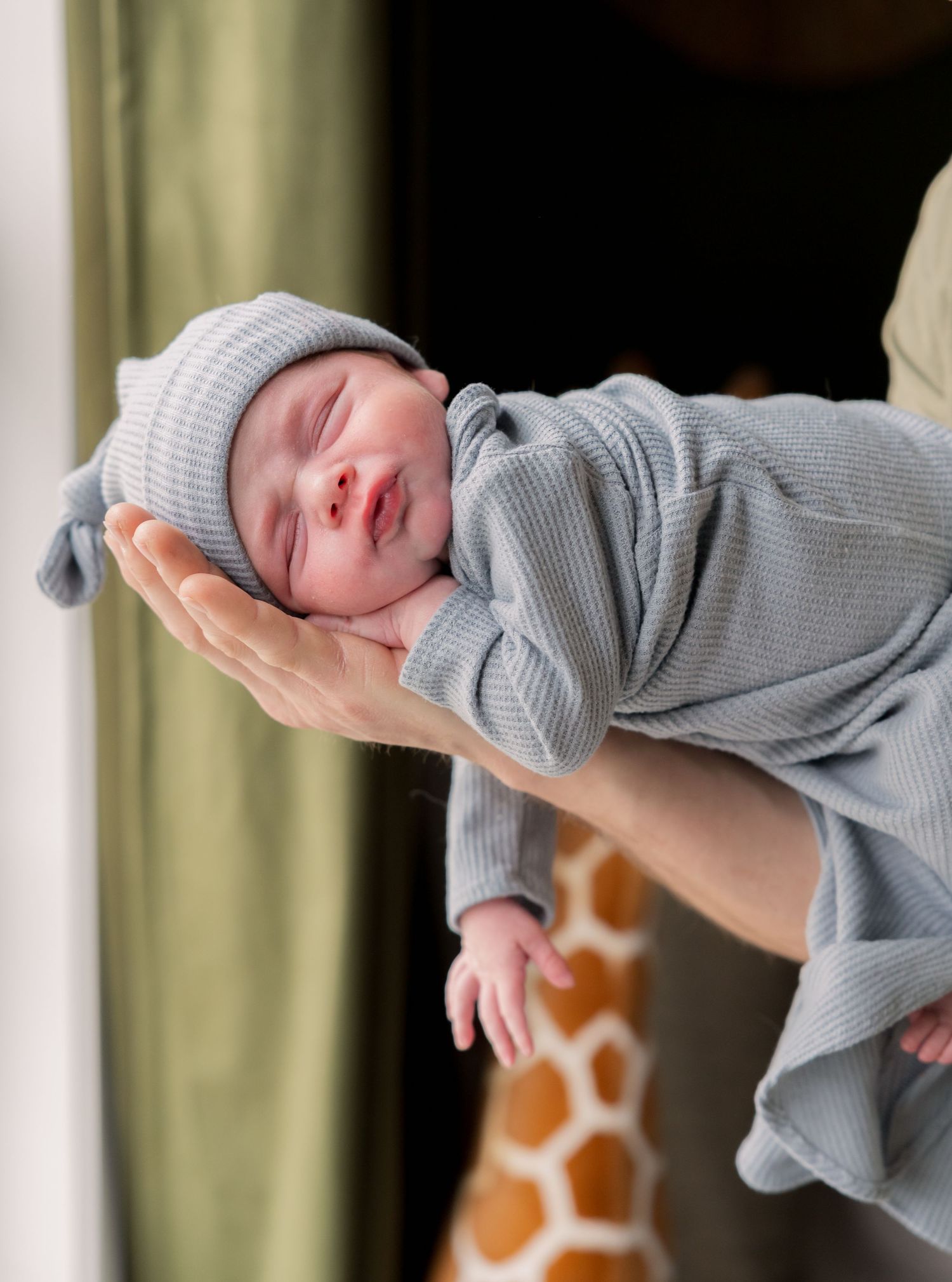 Newborn baby in gray knit outfit and hat cradled in adult hands, with a giraffe toy in the background.
