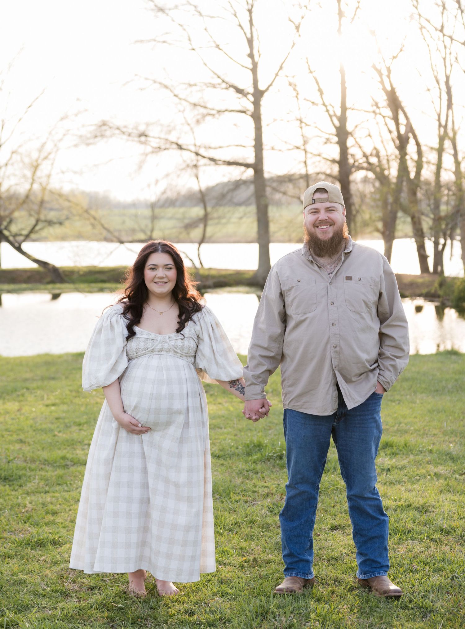 Pregnant couple holding hands outdoors near a lake, woman in plaid maternity dress, man in tan shirt and jeans, golden hour.