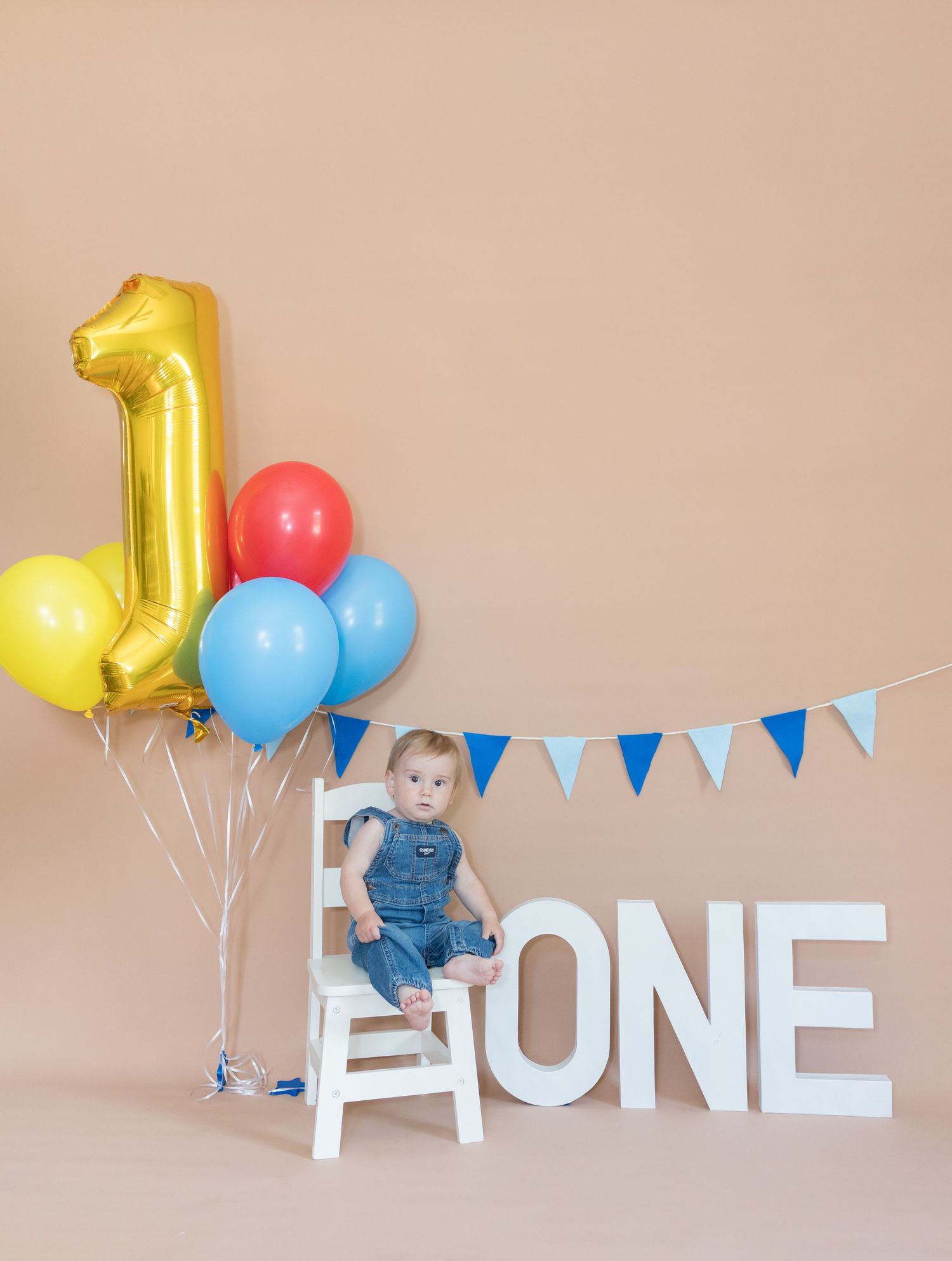Baby's first birthday photo with gold number one balloon, colorful balloons, blue bunting, and large ONE letters.