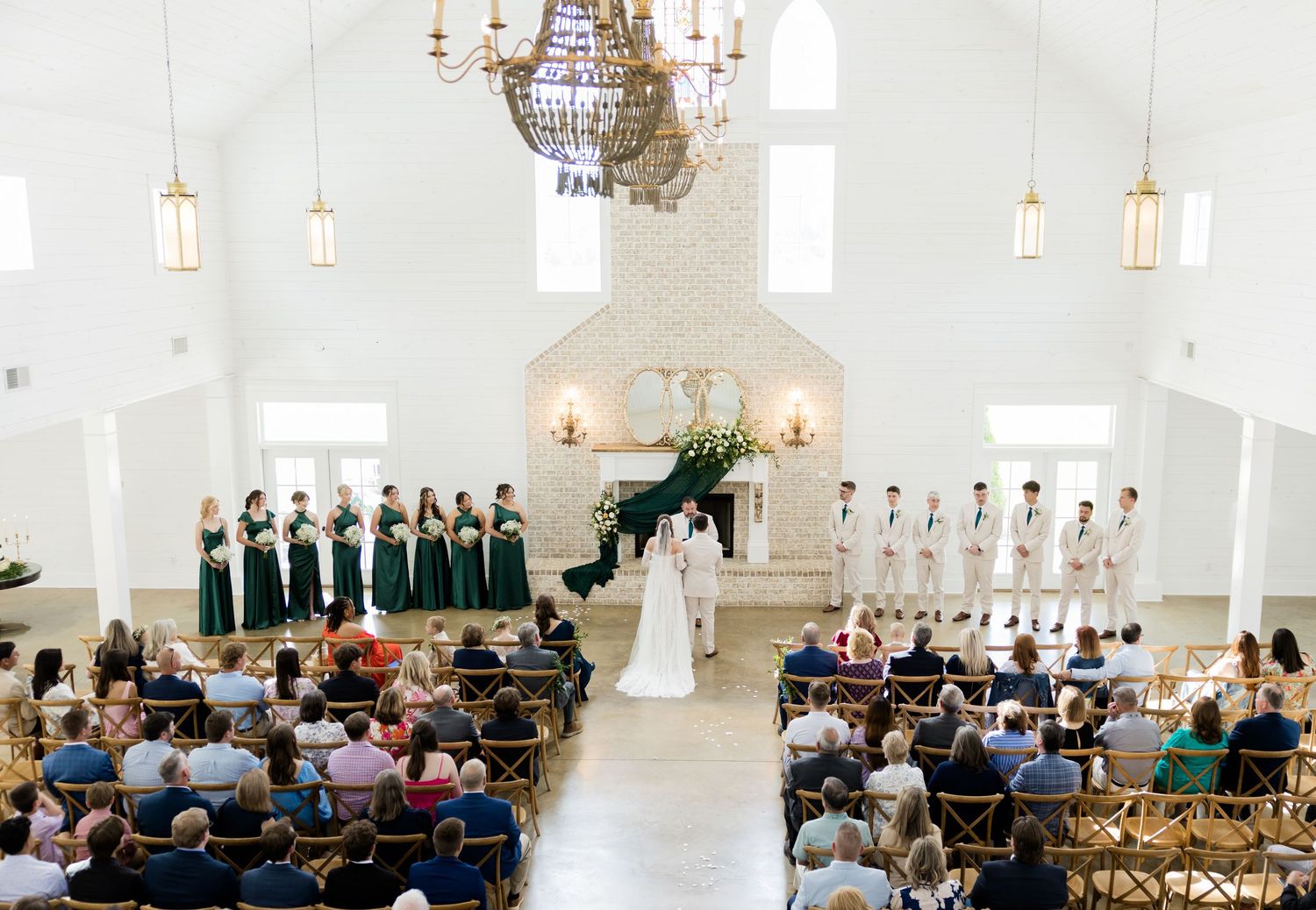 Elegant wedding ceremony in a white chapel with green bridesmaid dresses, floral arch, chandelier, and seated guests.