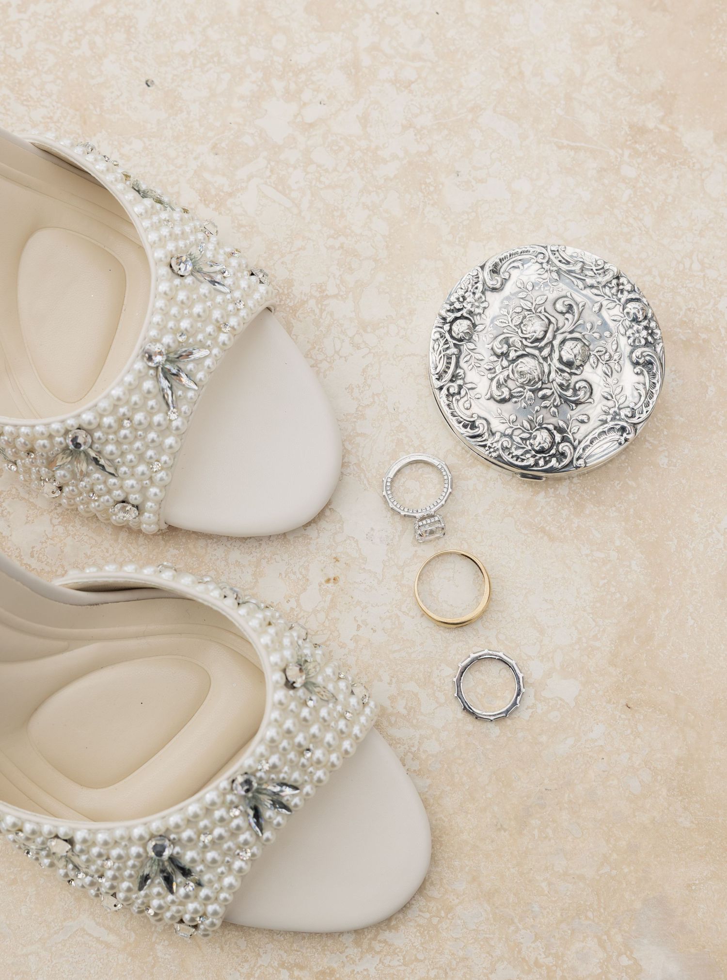 Pearl-embellished bridal flats displayed beside ornate silver compact and three wedding rings on marble surface.