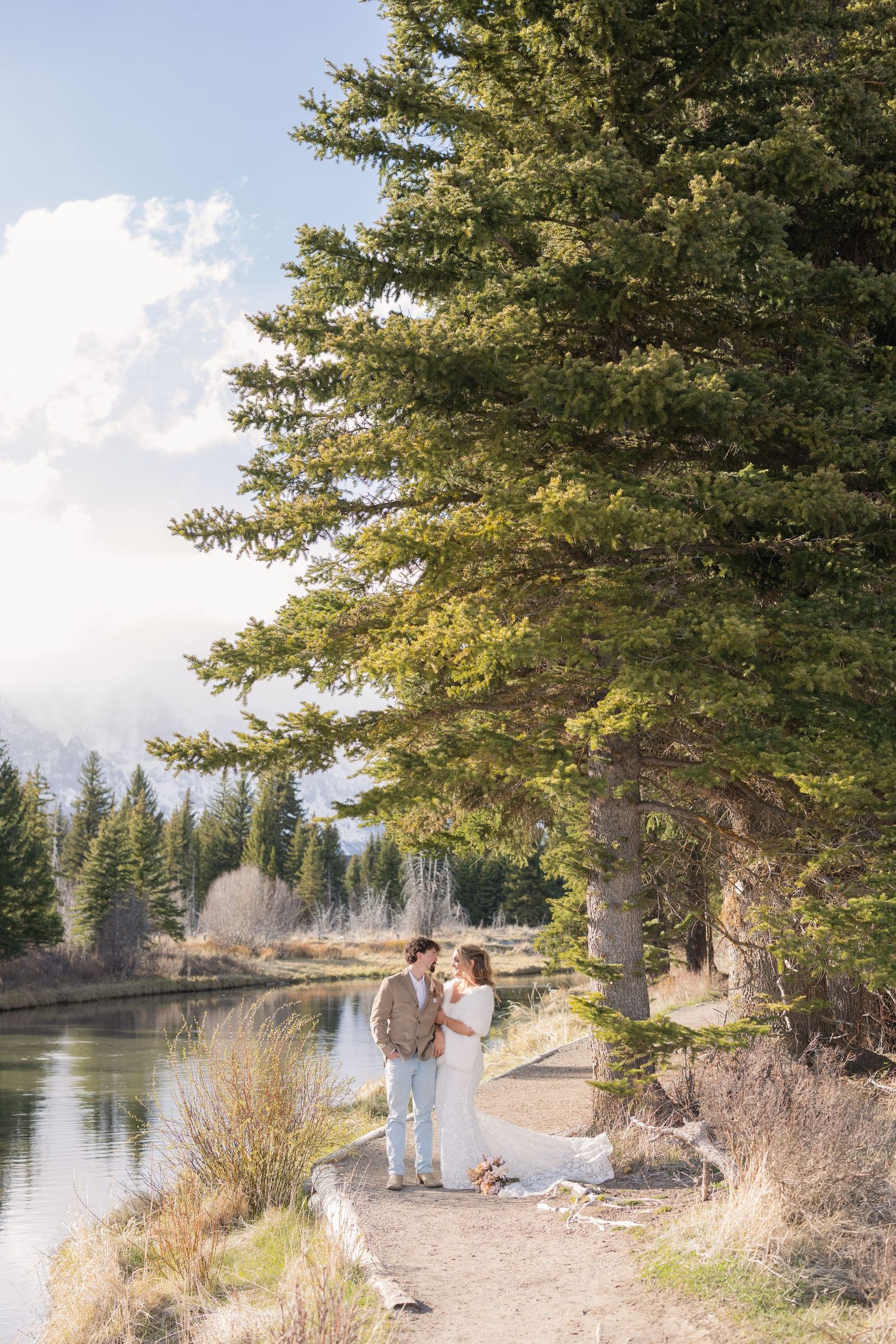 Couple shares a romantic moment by a mountain lake surrounded by tall evergreen trees in a scenic outdoor setting.