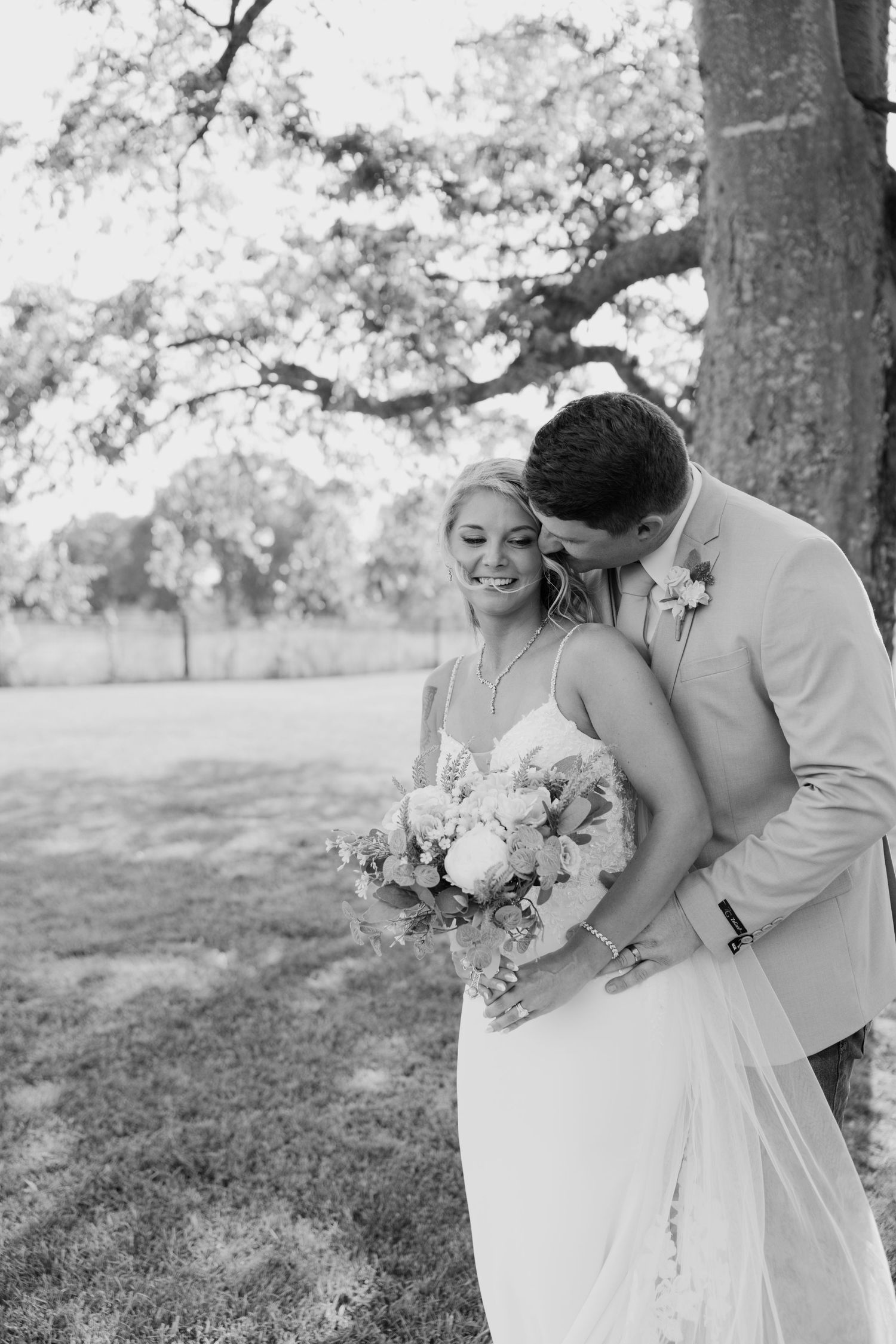 Black and white photo of a smiling bride holding a bouquet as the groom kisses her cheek outdoors near a large tree.