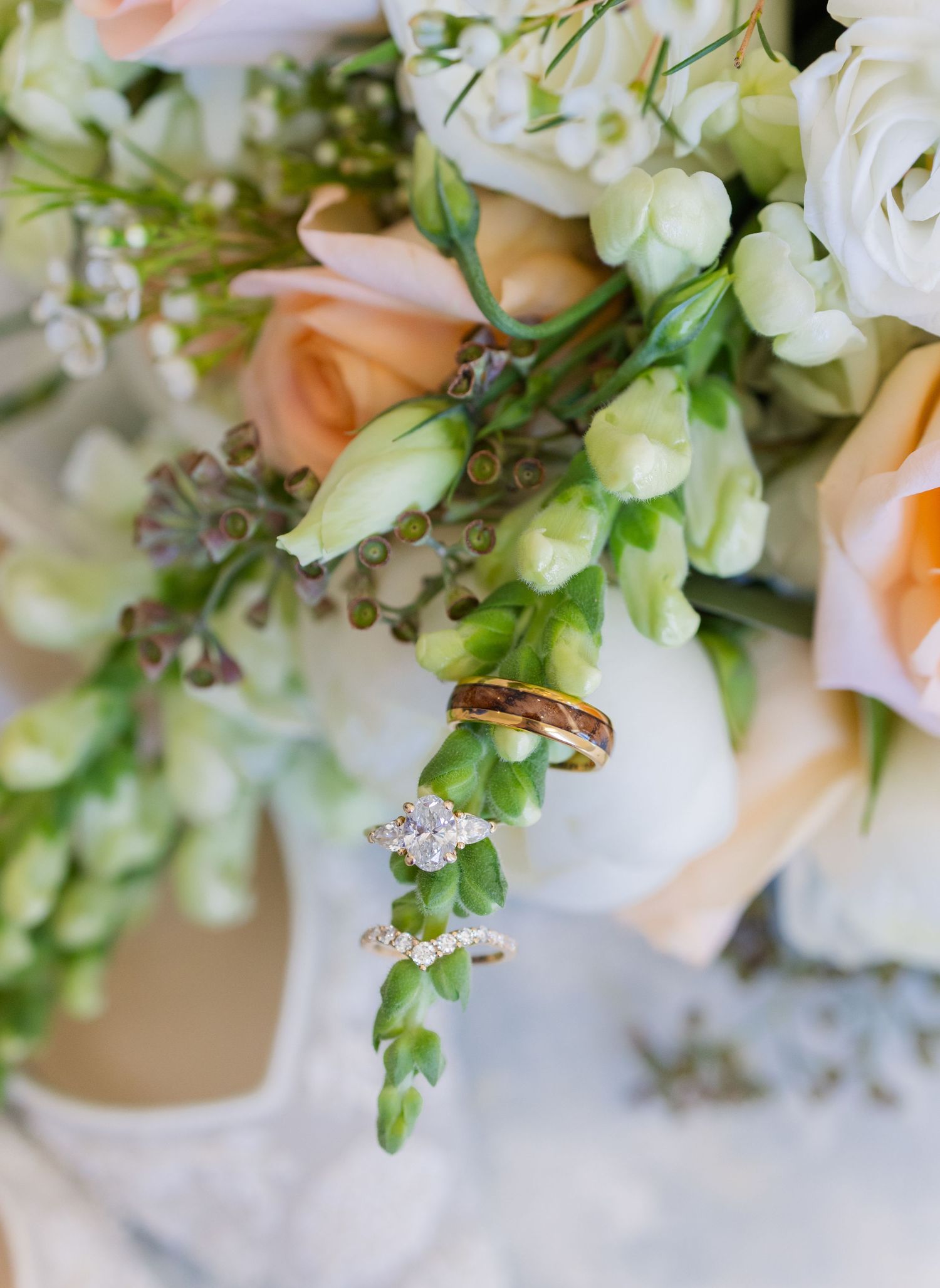 Wedding rings and diamond engagement ring nestled among peach roses and green flowers in a bridal bouquet.