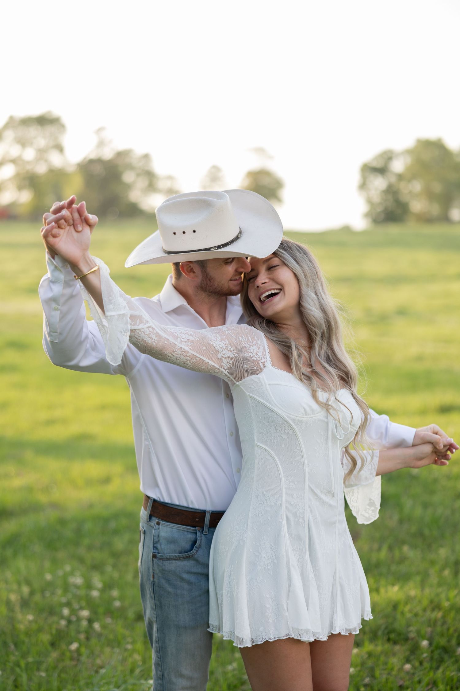 Couple dancing in a green field, man in white cowboy hat and shirt, woman in white lace mini dress, golden hour.