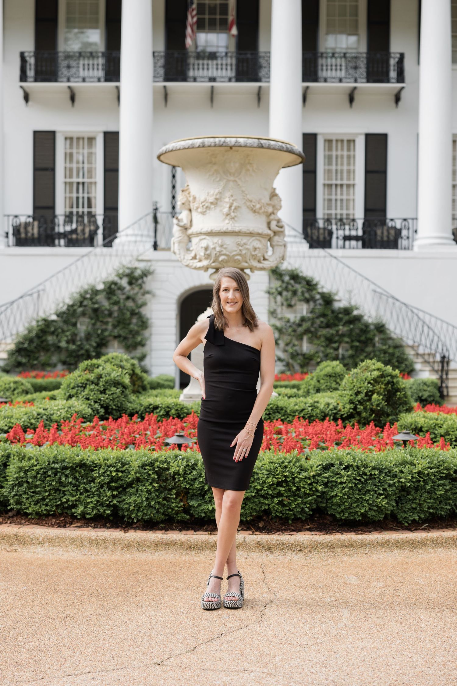 Woman in black one-shoulder dress poses in front of elegant white Southern mansion with red flower gardens at University Of Alabama for grad