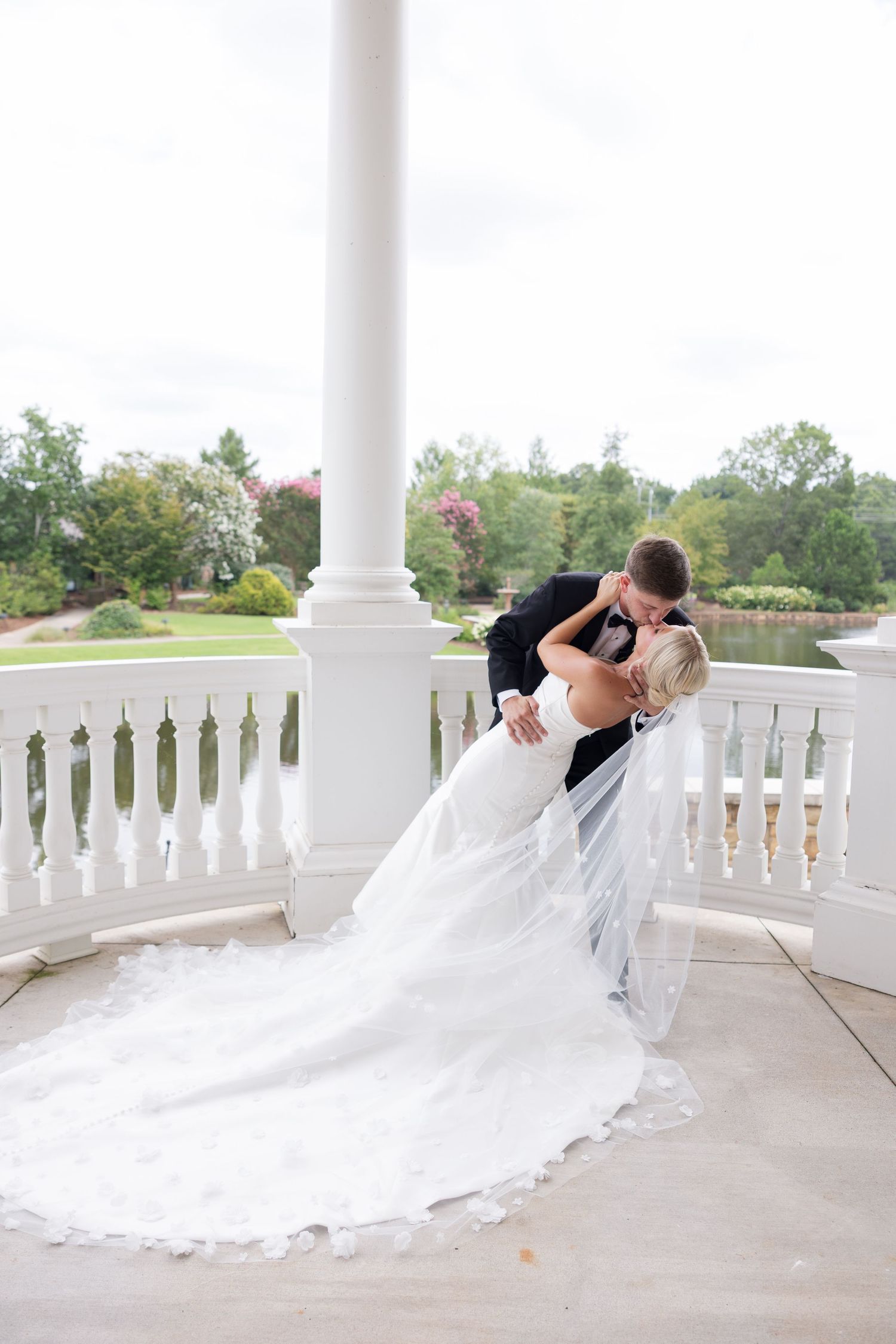 Bride and groom share a romantic dip kiss on a white balcony with lush garden views on their wedding day.