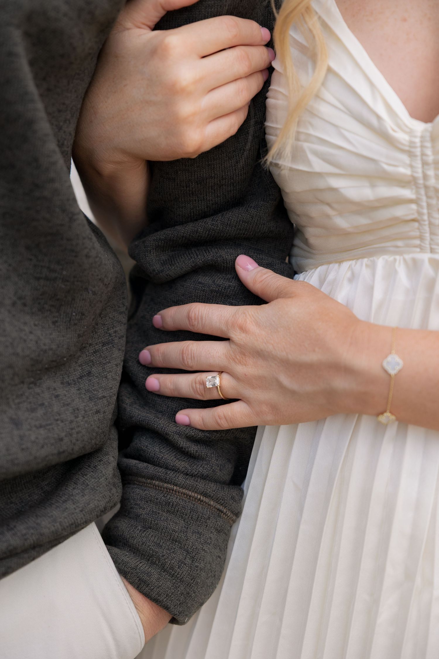 Close-up of woman's hands with engagement ring and bracelet holding man's arm in gray sweater.