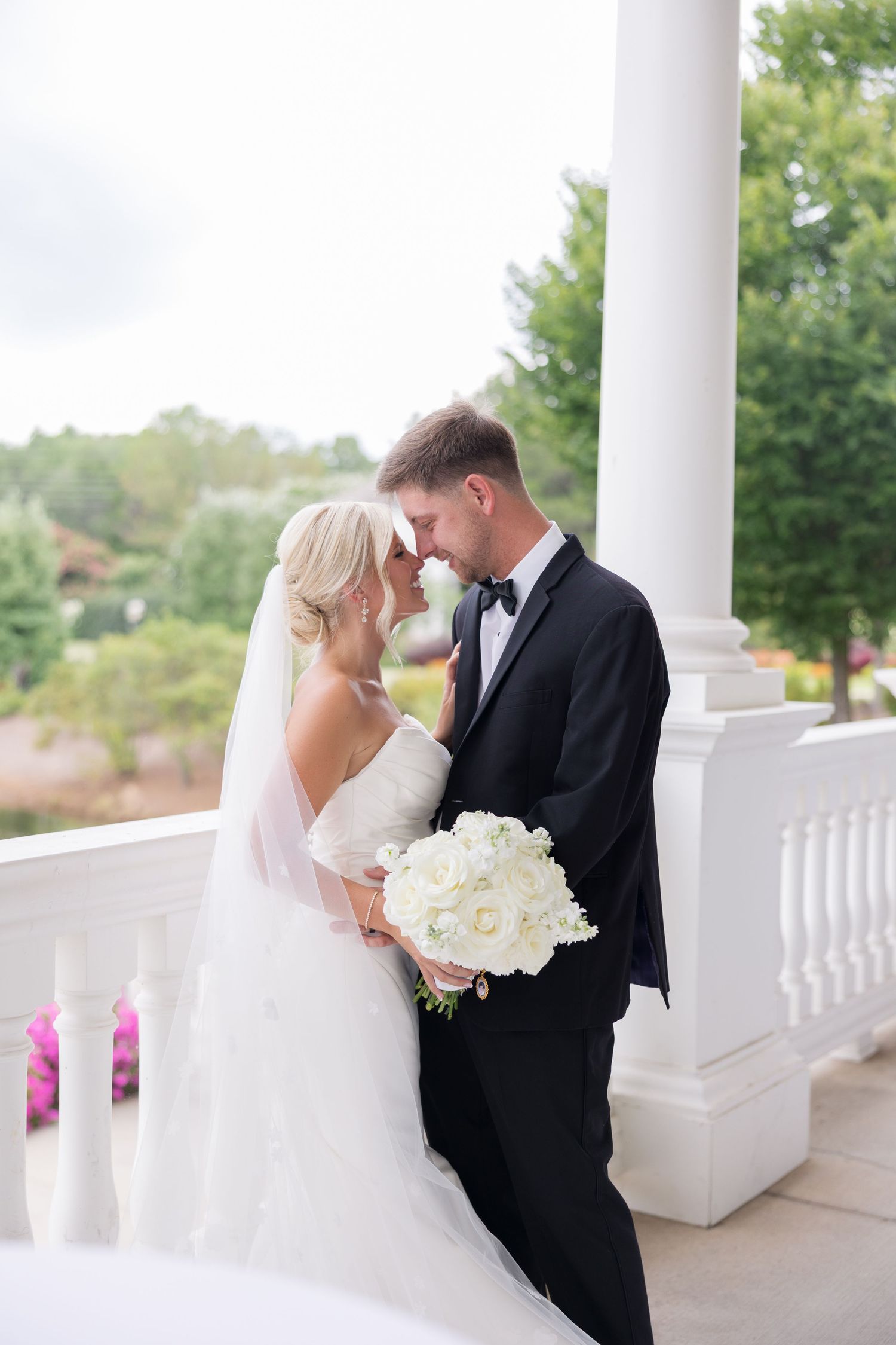 Bride and groom share an intimate forehead touch on a white columned veranda with lush greenery backdrop.