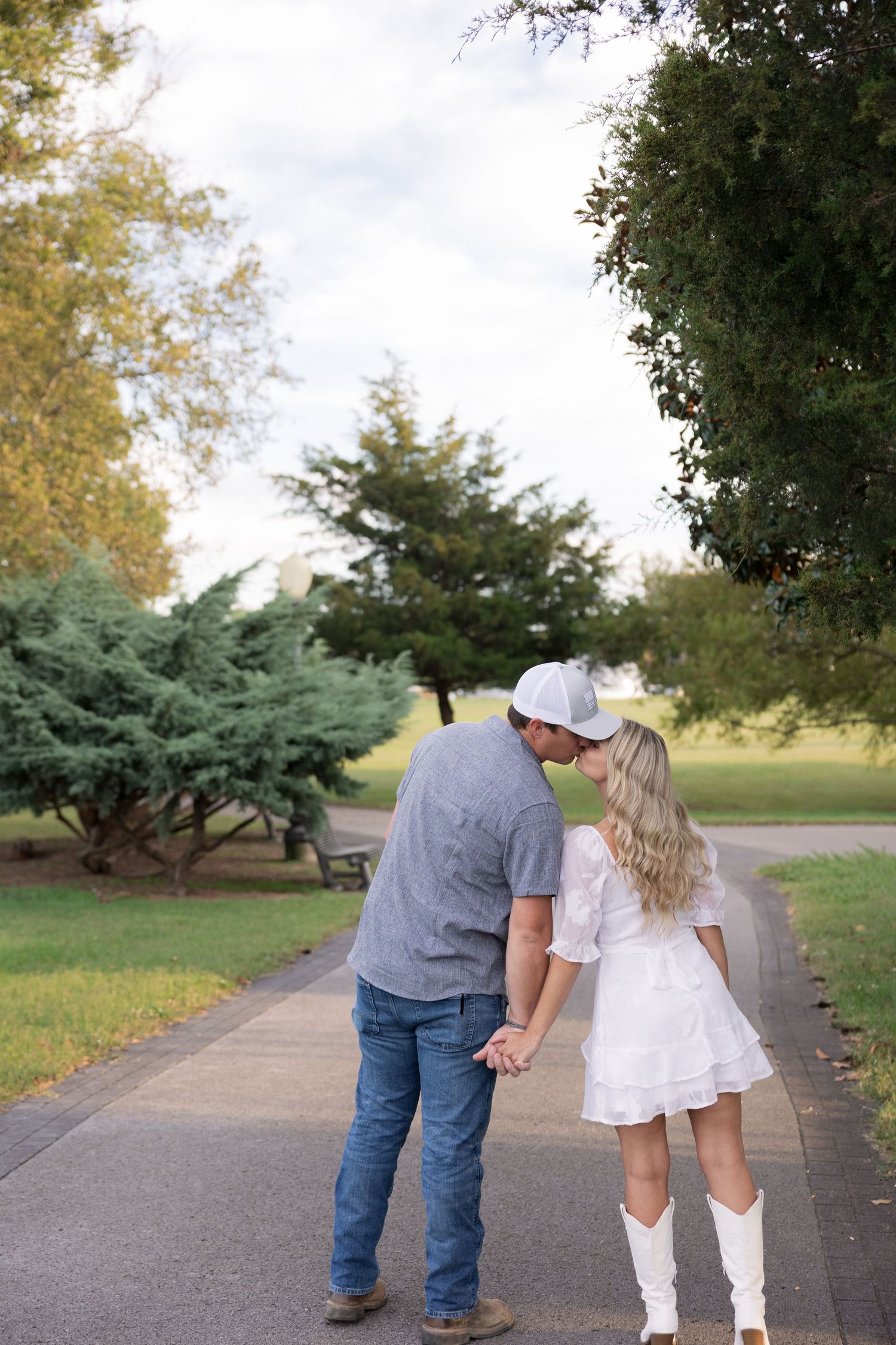 Couple sharing a kiss while holding hands on a park path, woman in white dress and boots, man in jeans and gray shirt.