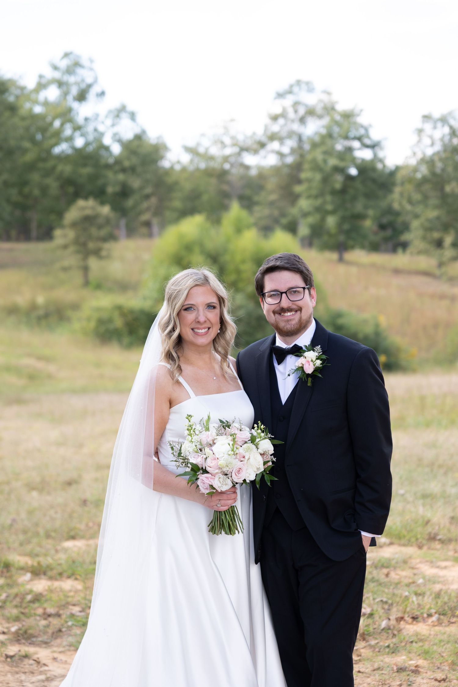 Smiling bride and groom pose outdoors in wedding attire, she holds a pink bouquet, he wears a black tuxedo with bow tie.
