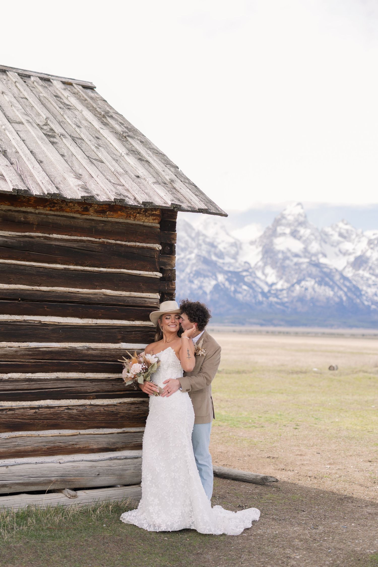 Bride and groom embrace beside a rustic log cabin with snow-capped Grand Teton mountains in the background.