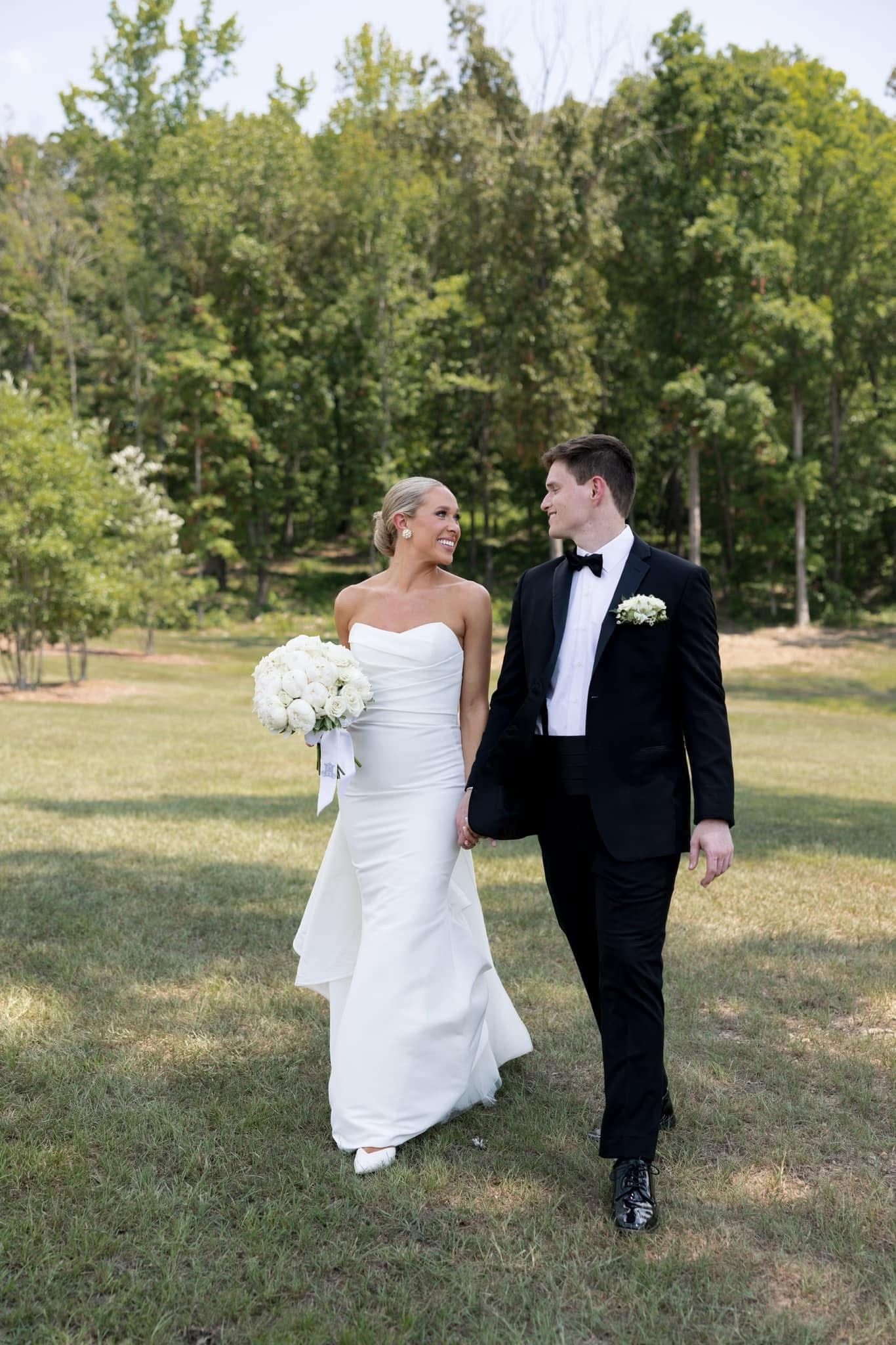 A bride and groom walk hand-in-hand across a green lawn, smiling at each other, she in a white gown, he in a black tuxedo.
