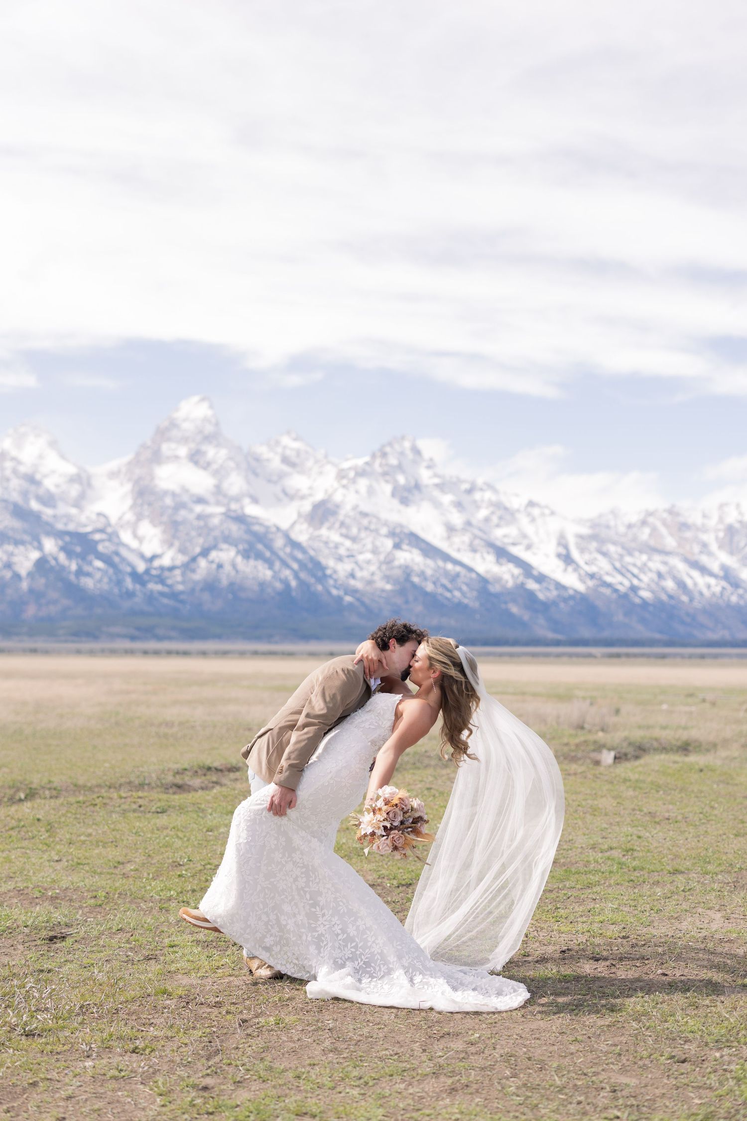 A bride and groom share a romantic dip kiss in a meadow with snow-capped Grand Teton mountains in the background.