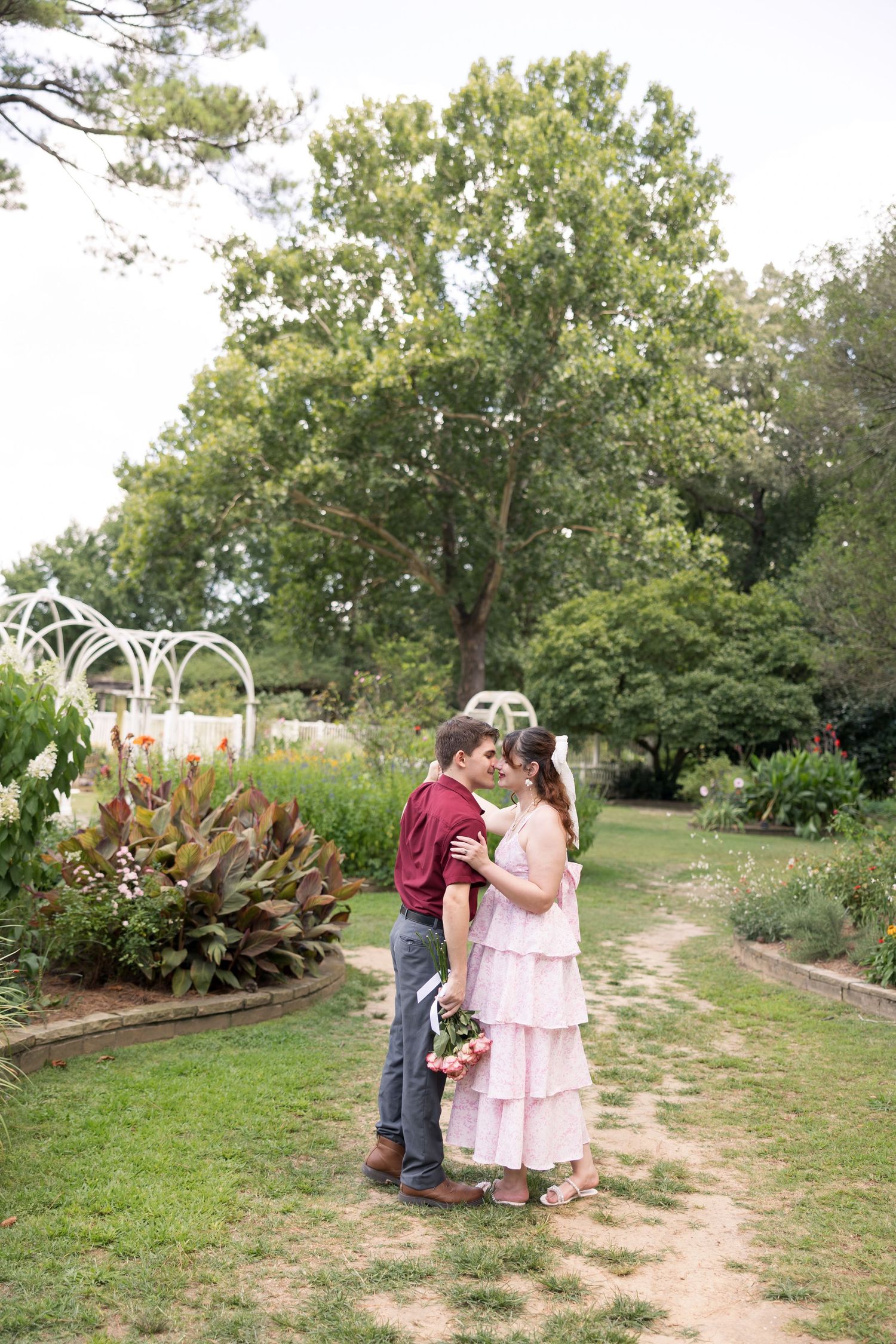 Couple sharing a romantic moment in a lush garden, woman in pink dress holding flowers, man in burgundy shirt.
