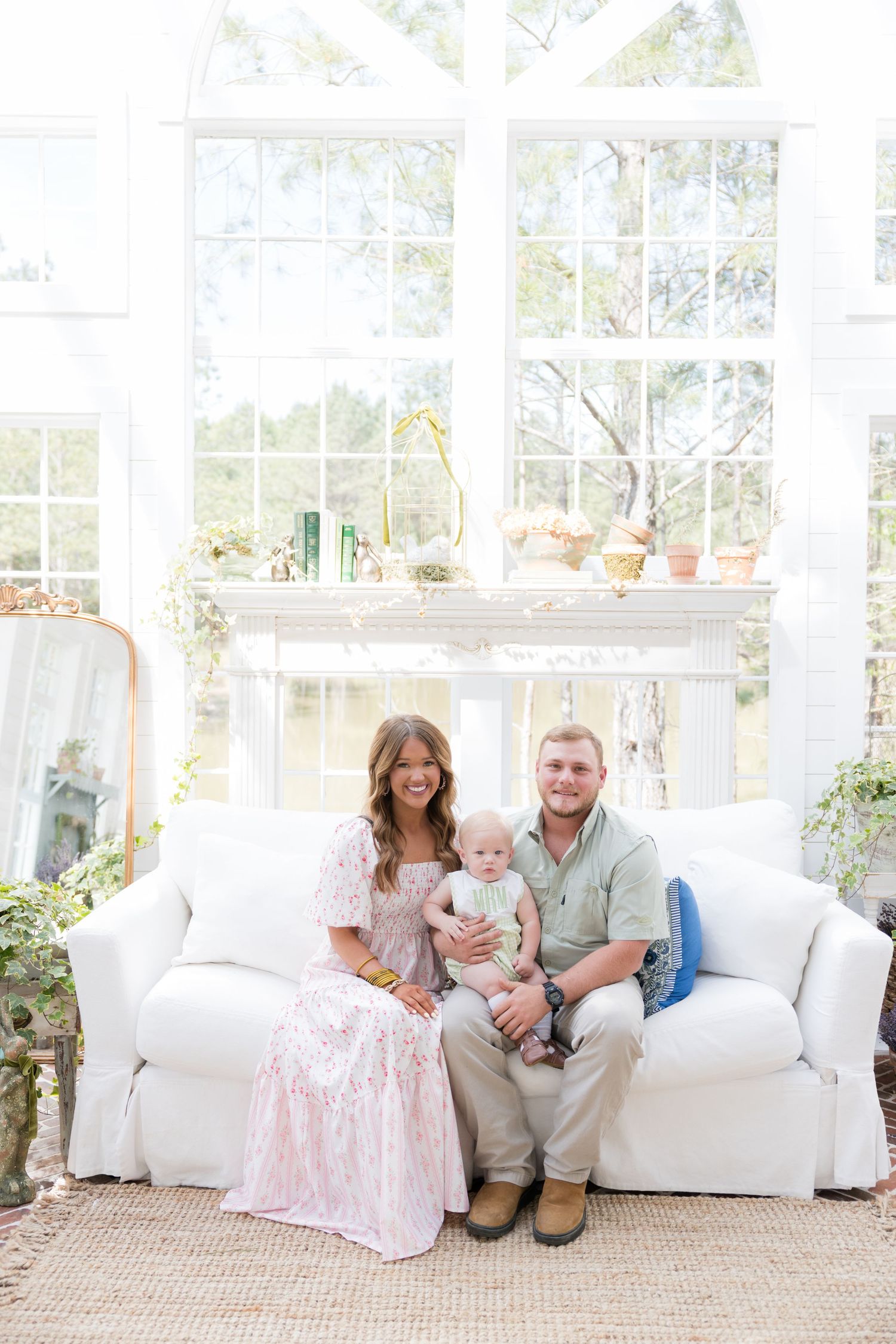 Young family with baby sits on white sofa in bright sunroom with large windows.
