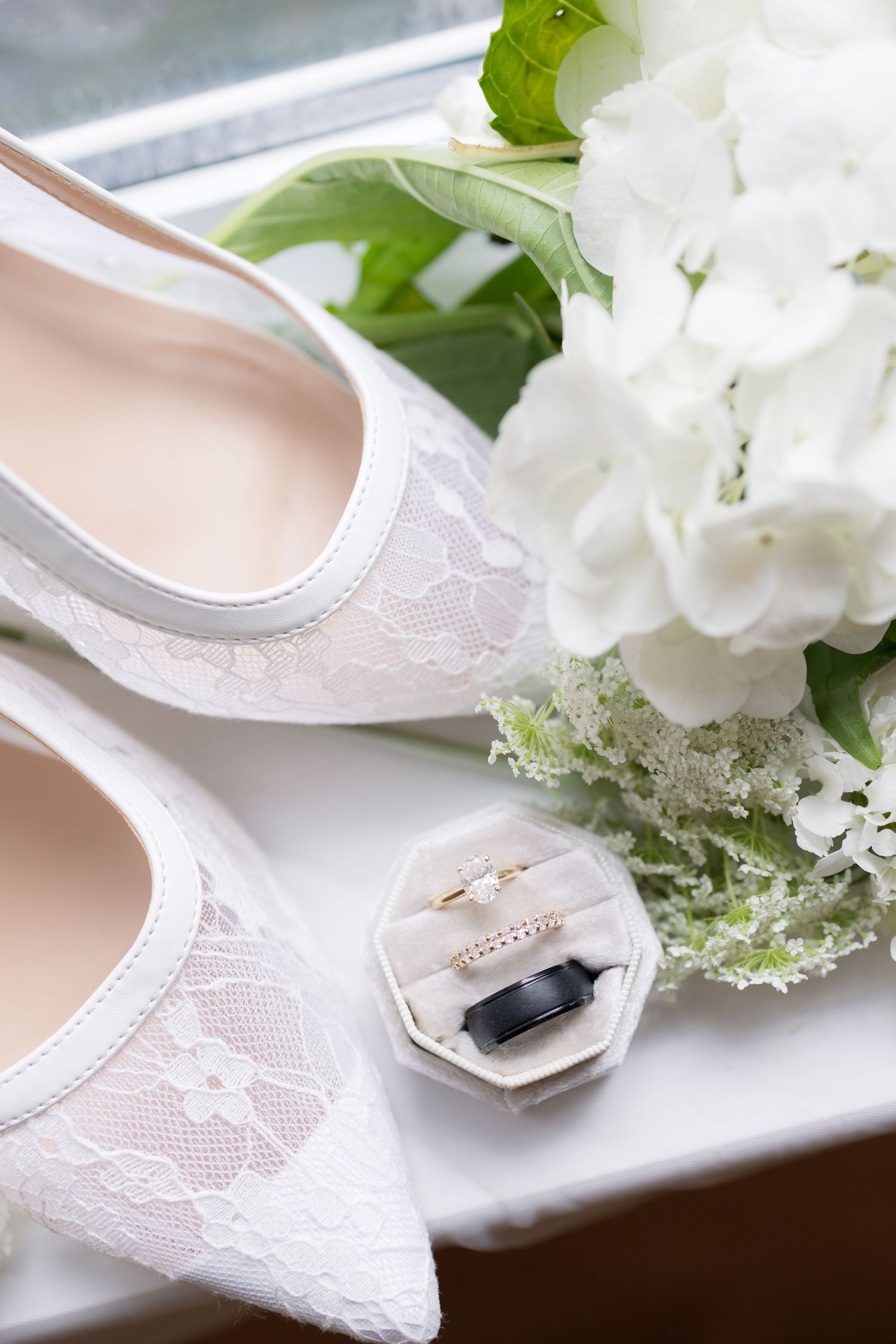 White lace bridal heels beside wedding rings in a box and white hydrangea bouquet on a windowsill.