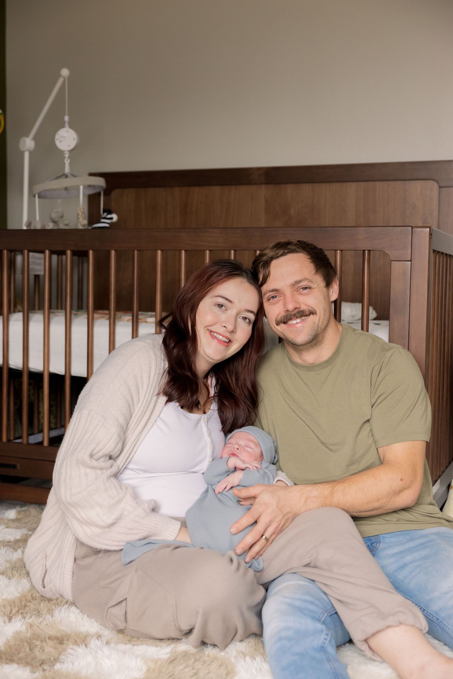 Happy couple sitting on floor holding newborn baby in front of wooden crib in nursery room.