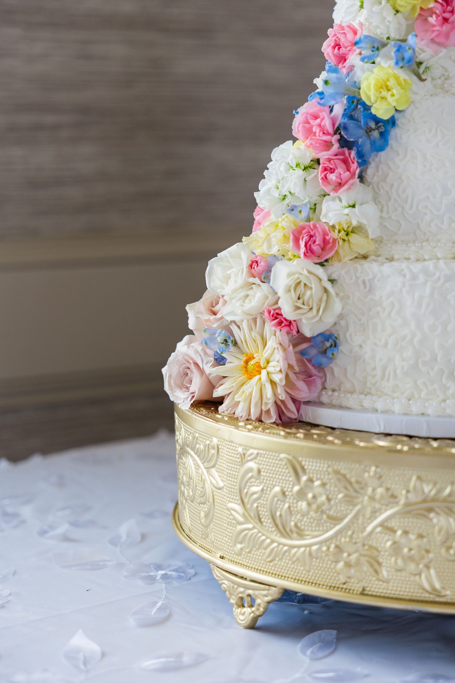 Elegant three-tier white wedding cake adorned with colorful fresh flowers on a gold ornate cake stand.