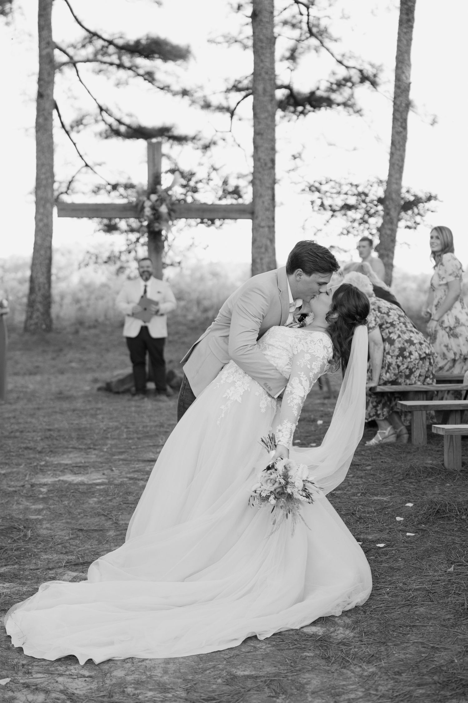 Black and white photo of groom dipping bride for a kiss at outdoor wedding ceremony surrounded by tall pine trees.