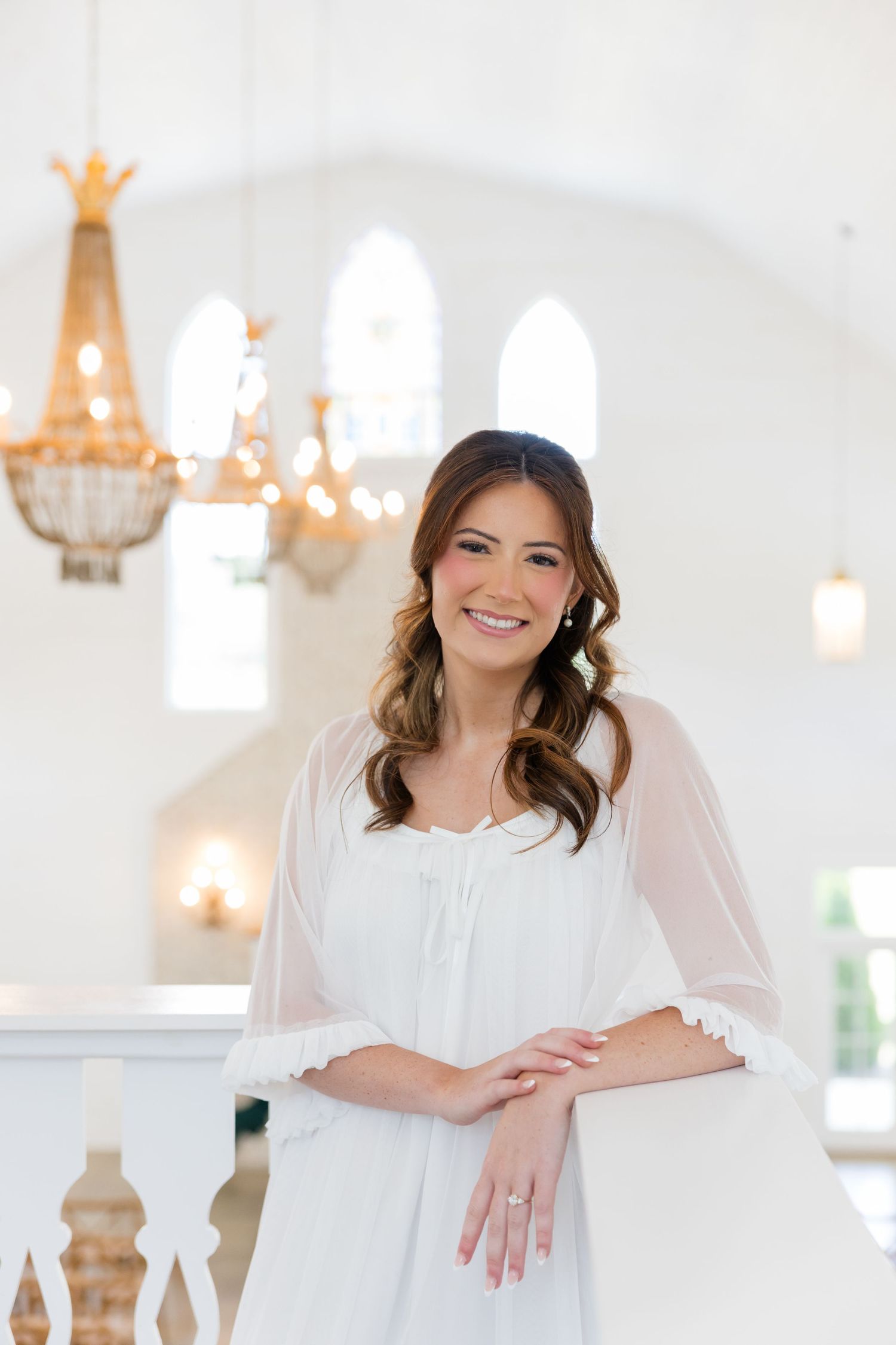 Smiling bride in a white chiffon robe leans on a railing inside an elegant white chapel with gold chandeliers.