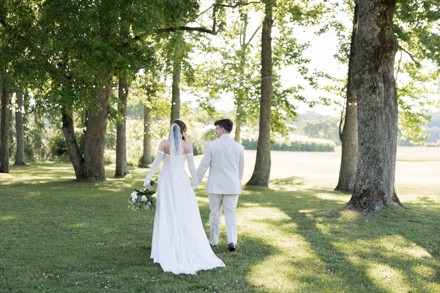 Bride and groom walk hand in hand across a green lawn surrounded by tall trees on their wedding day.