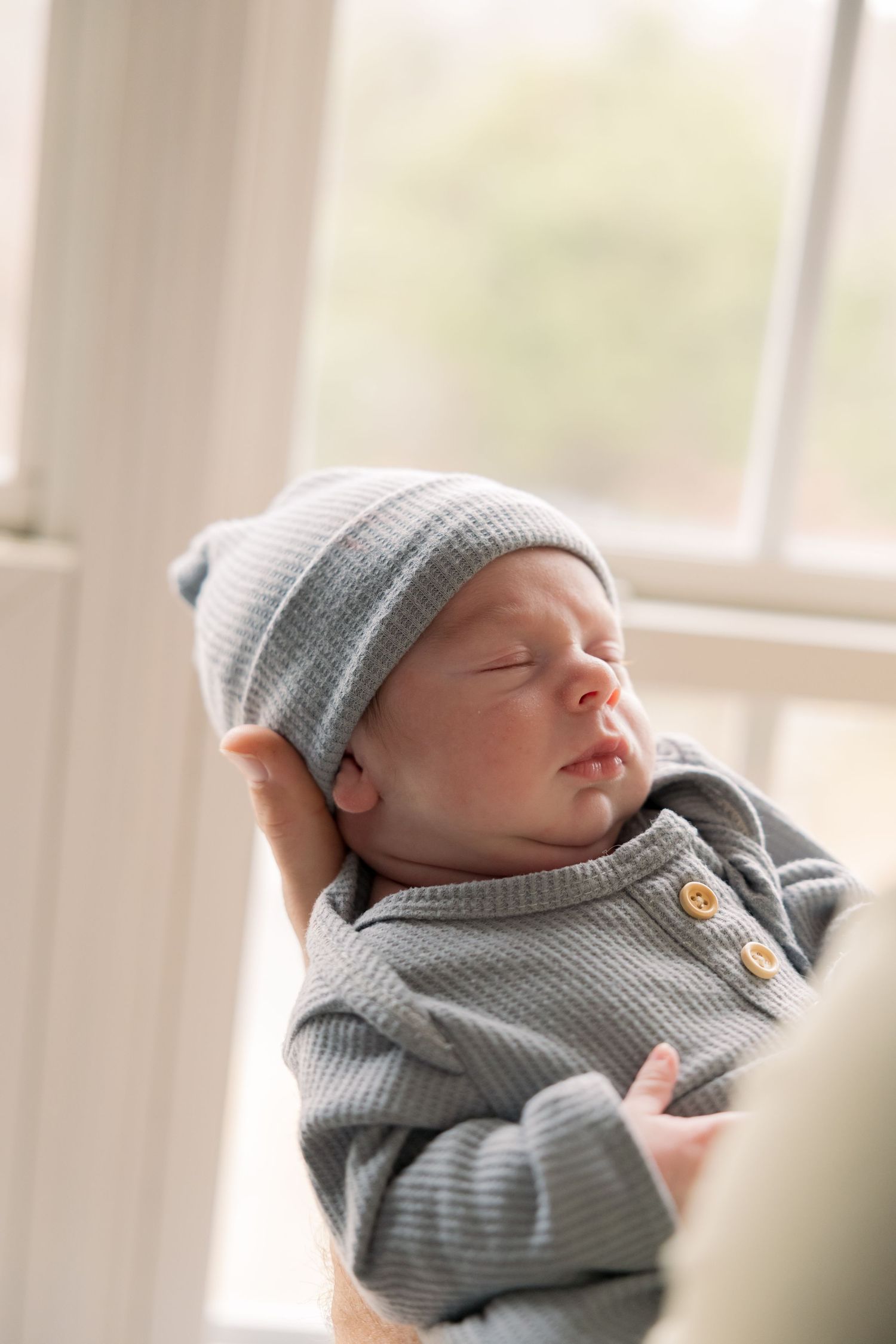 Newborn baby sleeping peacefully in gray knit outfit and matching beanie hat, cradled in loving arms near a bright window.