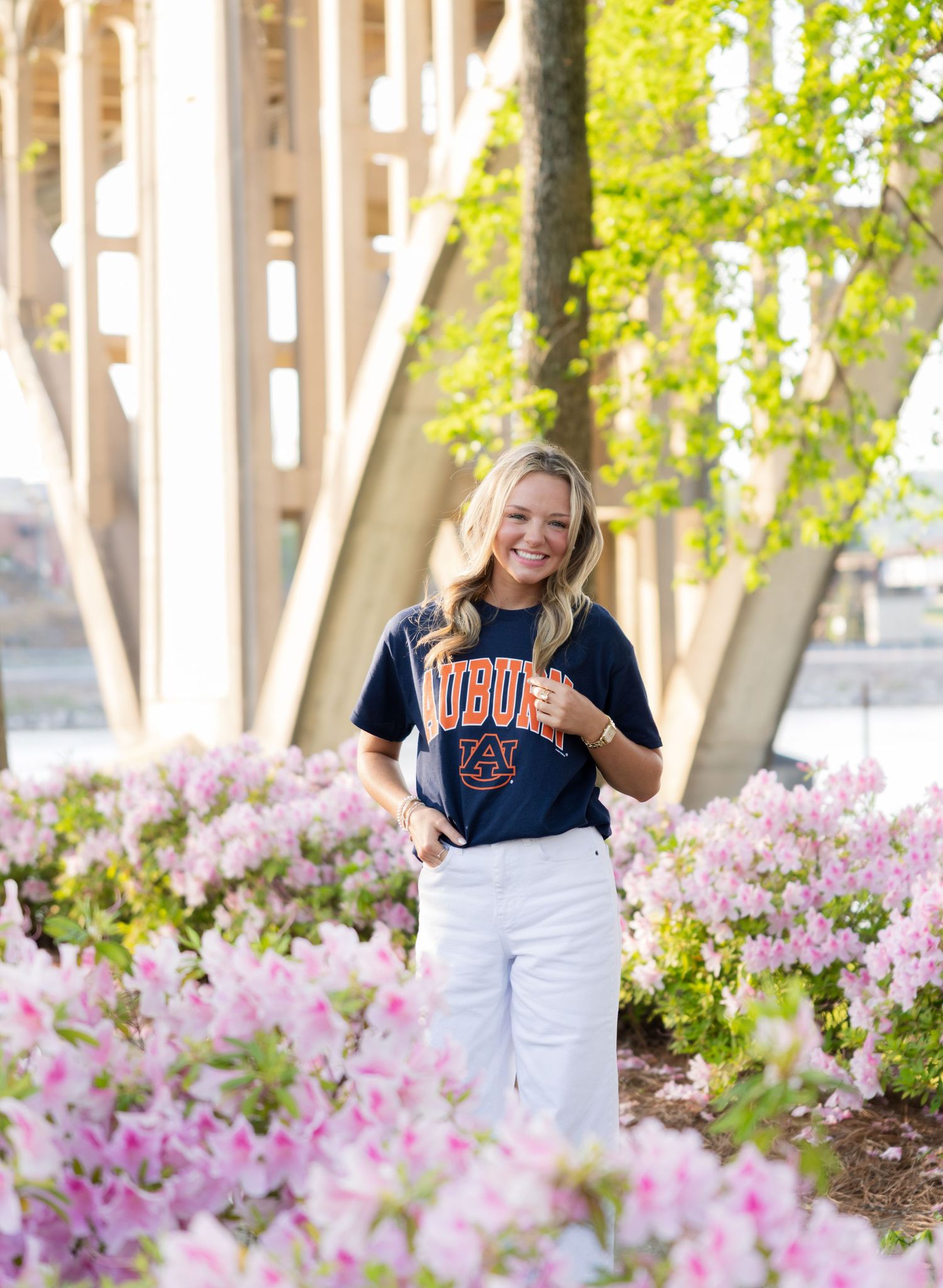 Smiling blonde woman in Auburn University navy t-shirt and white pants standing among pink azalea blooms outdoors.