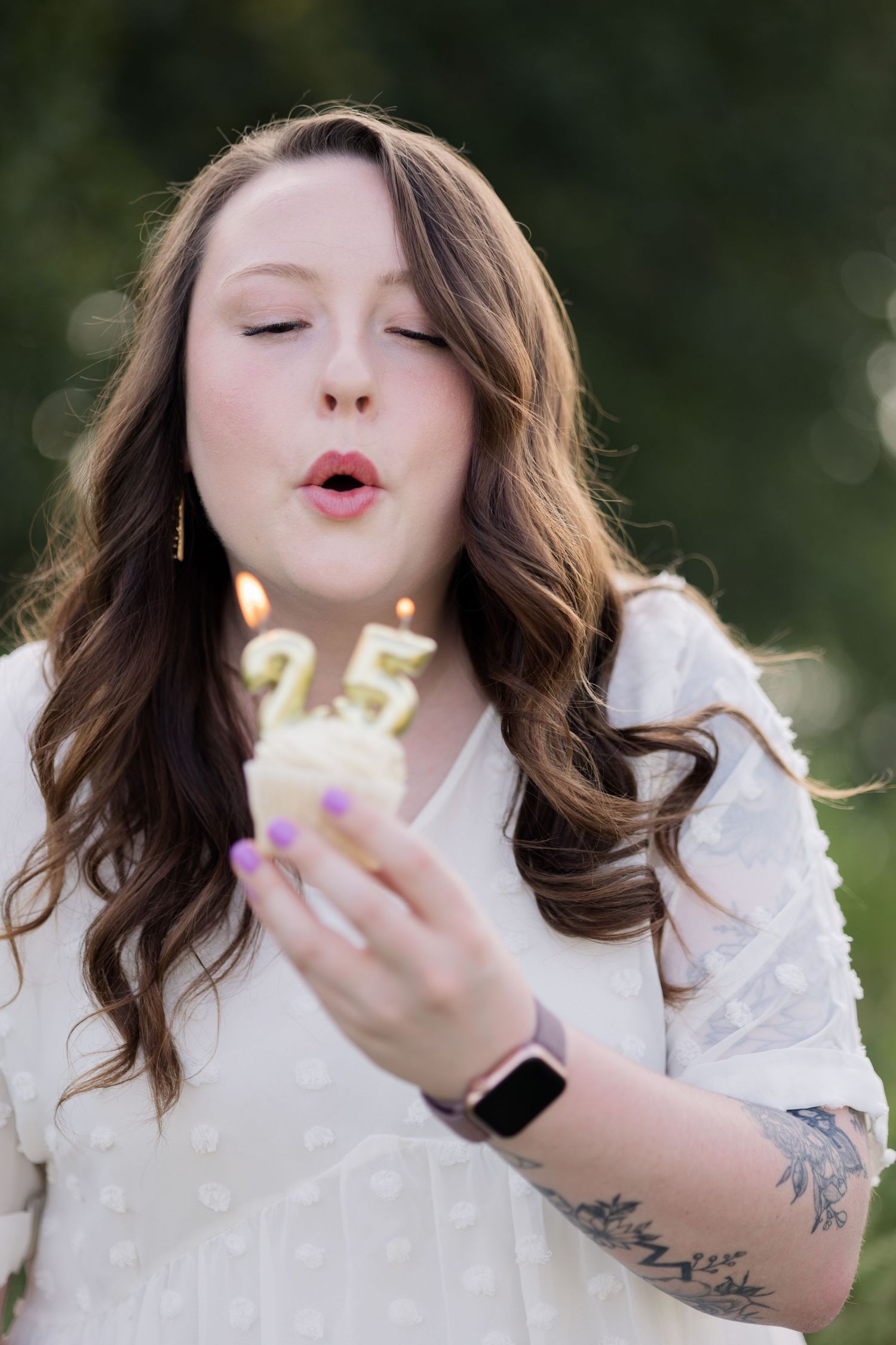 Woman blowing out number 25 candles on a cupcake, celebrating her 25th birthday outdoors.