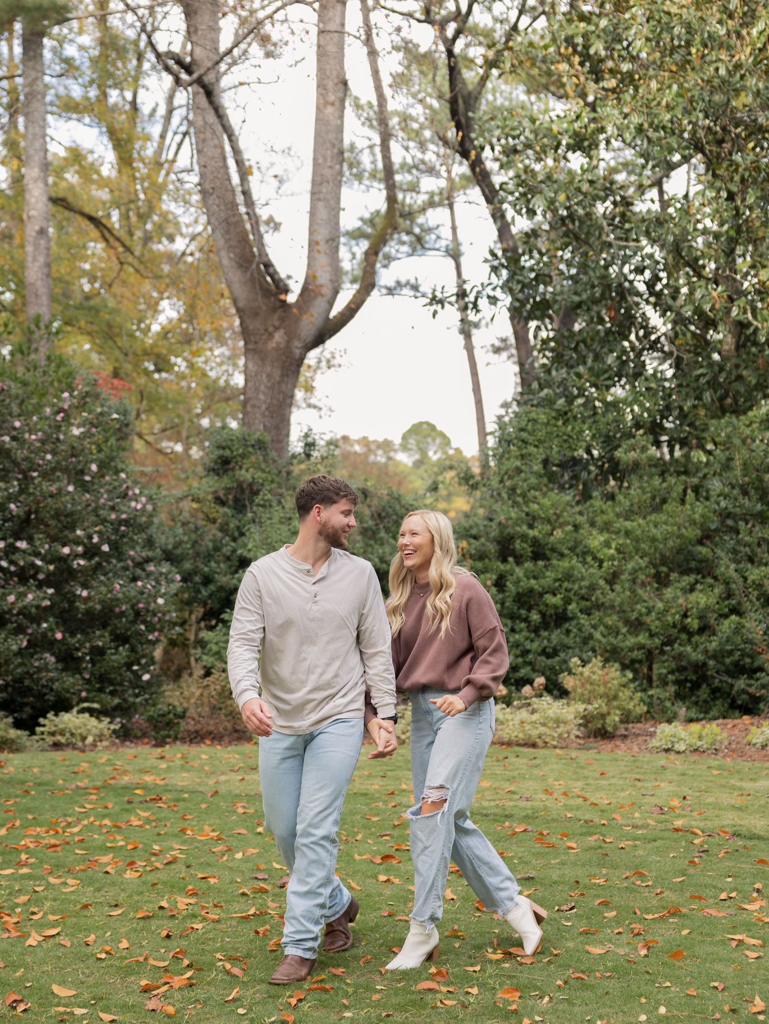 A couple holds hands and smiles at each other while walking through a lush green garden in autumn.