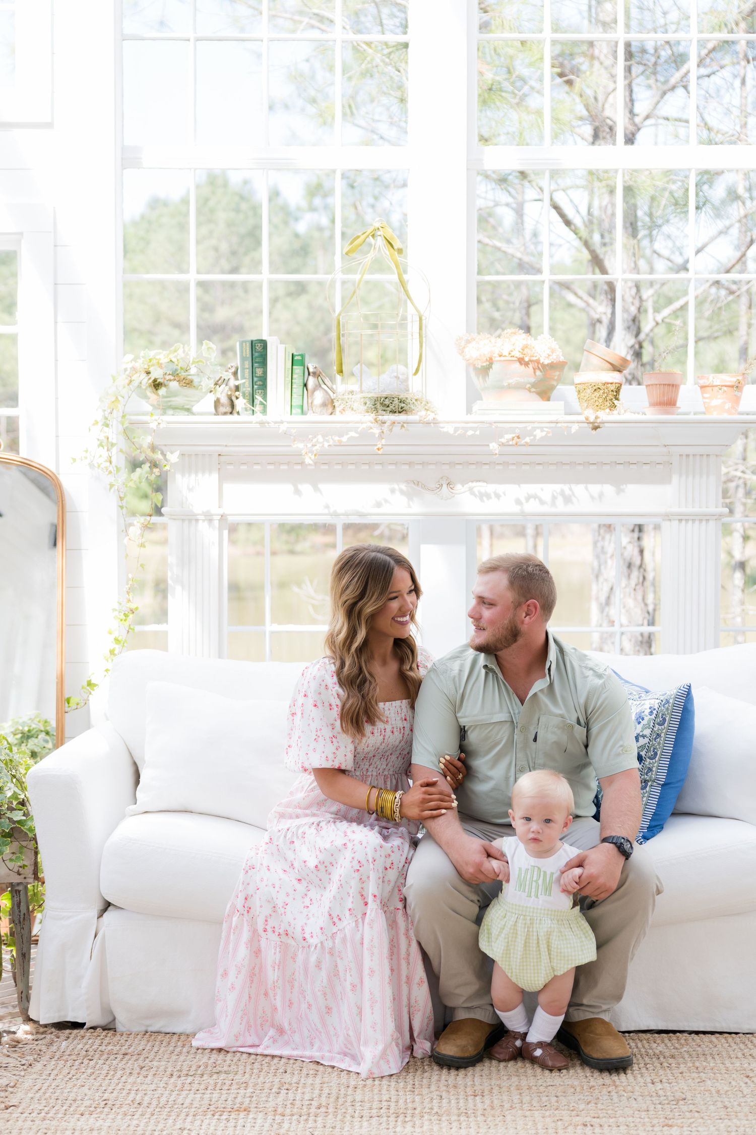 Family portrait of couple with baby on white sofa in bright sunroom with fireplace and large windows.