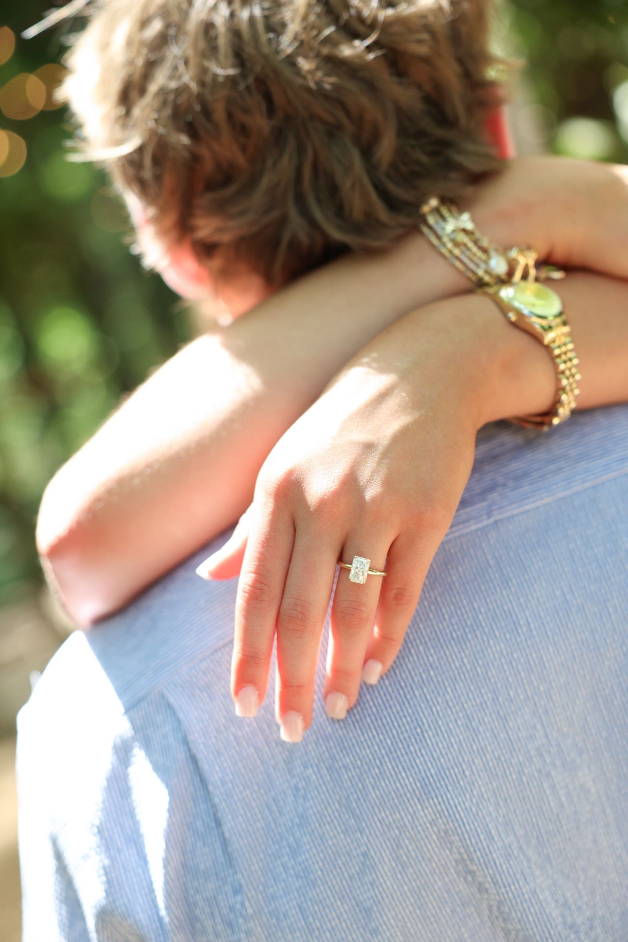 Woman showing off diamond engagement ring while embracing partner outdoors in sunlight.