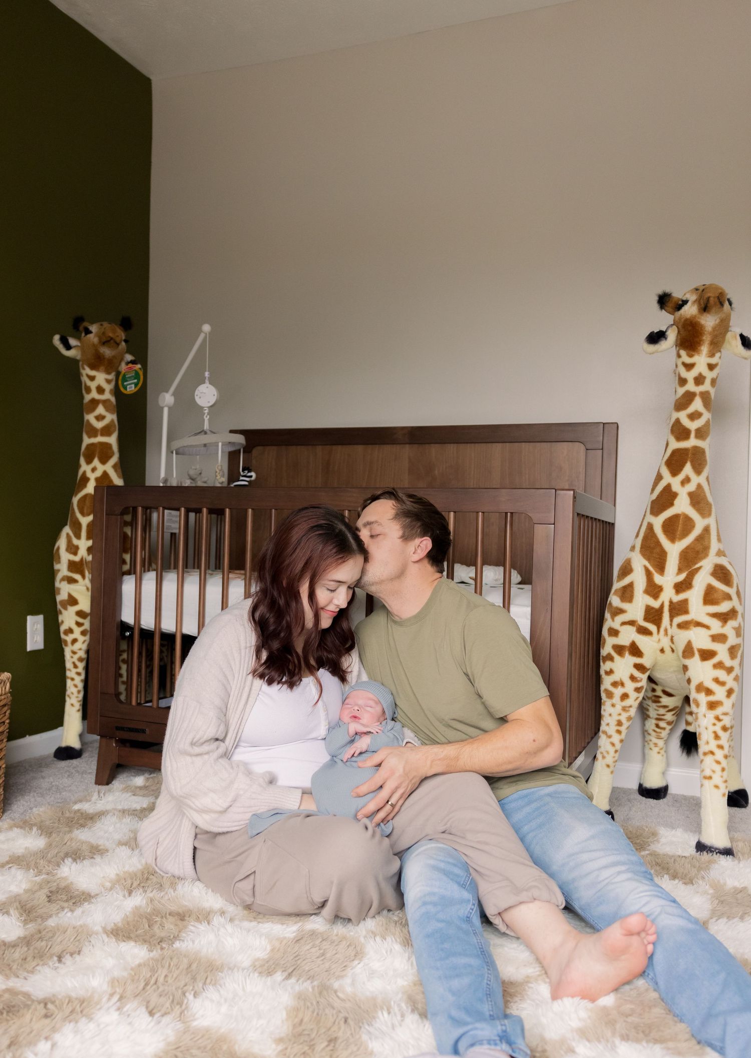 New parents sitting on nursery floor with newborn, surrounded by giraffe decor and wooden crib.
