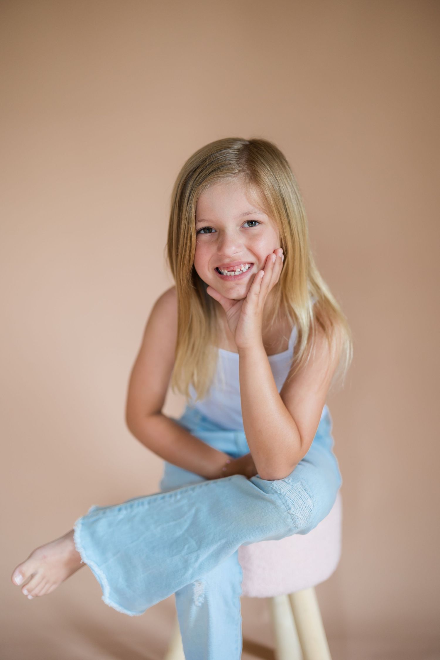 Smiling young girl with blonde hair sitting on a stool, wearing a white top and blue pants against a beige background.